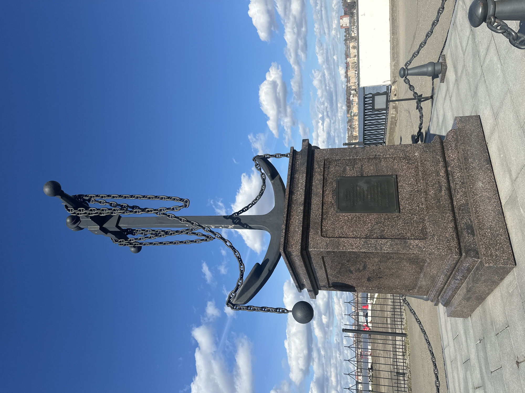 The image depicts a monument in Votkinsk, Russia, featuring a dark-colored anchor mounted on a granite pedestal. The anchor is adorned with chains and spheres. A plaque is visible on the pedestal. The monument stands on a paved area, with a metal fence and a cityscape visible in the background. The sky is blue with scattered white clouds.