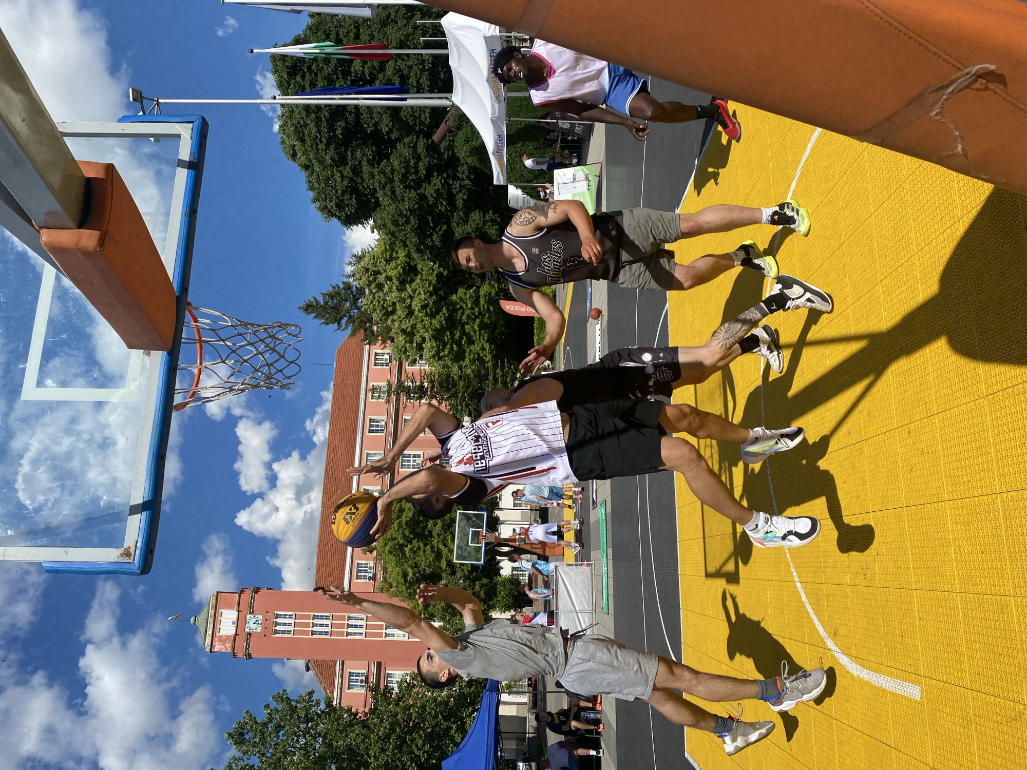 The image captures an outdoor street basketball game taking place in the city of Pleven, Bulgaria. In the foreground, four men are actively engaged in the game. Two players are jumping for a rebound or a shot near the basket, with one player in a white and red striped jersey holding the ball. Another player in a grey t-shirt and shorts is reaching up towards the ball. To the right, two other players are positioned on the court. The background reveals a characteristic Pleven building with a prominent red clock tower, suggesting the game is being played in a public space, possibly a square or park, within the city. Several tents and flags are also visible, indicating a possible event or organized competition. The sky is bright blue with scattered clouds, and the court is bright yellow, characteristic of specialized urban sports surfaces.