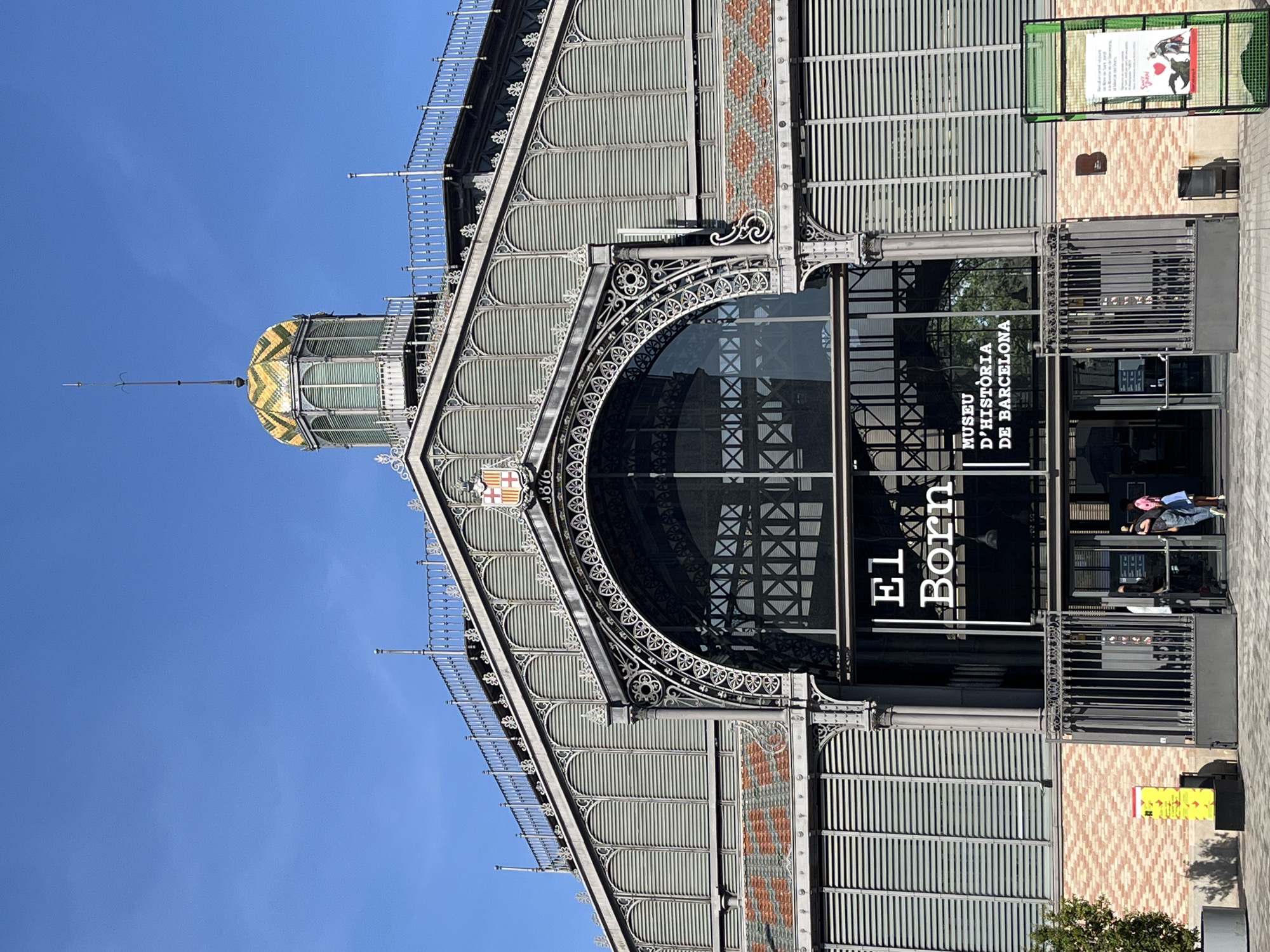 The image captures the grand, ornate iron and glass architecture of El Born Centre de Cultura i Memòria in Barcelona, Spain, under a clear blue sky. The historical building features a prominent arched entrance with large glass panels, reflecting parts of the exterior. Above the entrance, intricate grey metalwork forms decorative patterns and structural supports across the facade.
A distinctive green and yellow tiled dome or cupola, topped with a slender spire, is visible on the left side of the structure. The upper sections of the building are composed of corrugated or slatted panels, interspersed with decorative brickwork or tiles in a terracotta and light-colored diamond pattern. A coat of arms and the year "1876" are discernible on the upper left facade, indicating the building's heritage.
The main entrance prominently displays "EL BORN" in large white letters on the left glass panel and "MUSEU D'HISTÒRIA DE BARCELONA" (Museum of History of Barcelona) in smaller text on the right. Several casually dressed individuals are visible near the entrance, some seated and others standing, with one person having bright pink hair. The ground level in front of the building is paved and includes sections of patterned brickwork. A small green sign frame with a poster and a partially visible yellow sign are also present. The bright, clear sky suggests a sunny daytime setting.
