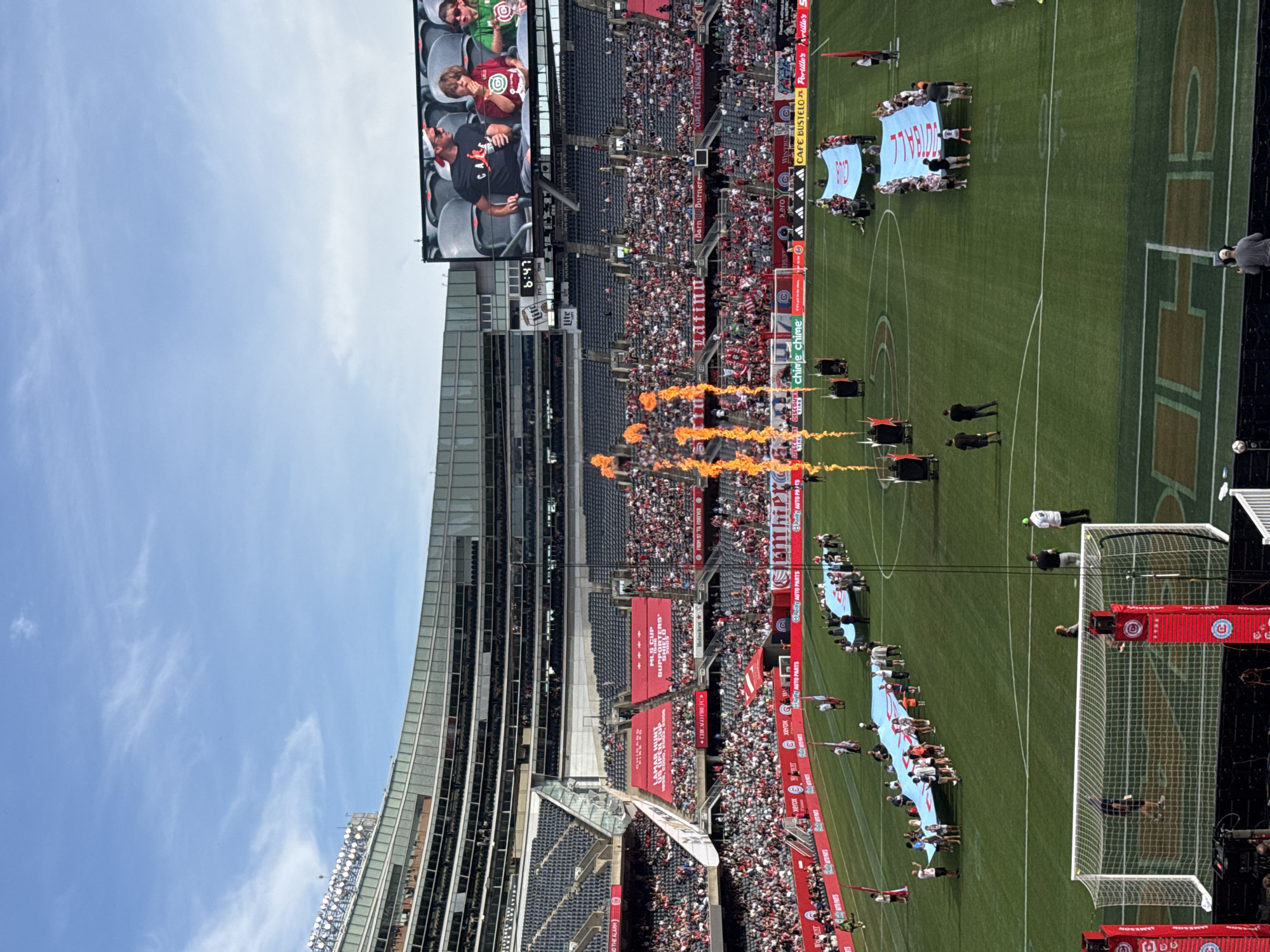 The image captures a daytime outdoor scene at Soldier Field in Chicago, Illinois, during a soccer match. The stadium, a prominent Chicago landmark, is filled with spectators in the stands, many wearing red. On the green soccer field, a large blue banner is being held by a group of people. Flames erupt from the field in the foreground, suggesting a pre-game ceremony or a dramatic moment in the match. A large video screen is visible in the background, displaying images of people. The sky above is partly cloudy, indicating fair weather. The presence of a soccer goal in the lower portion of the frame confirms the sporting event.