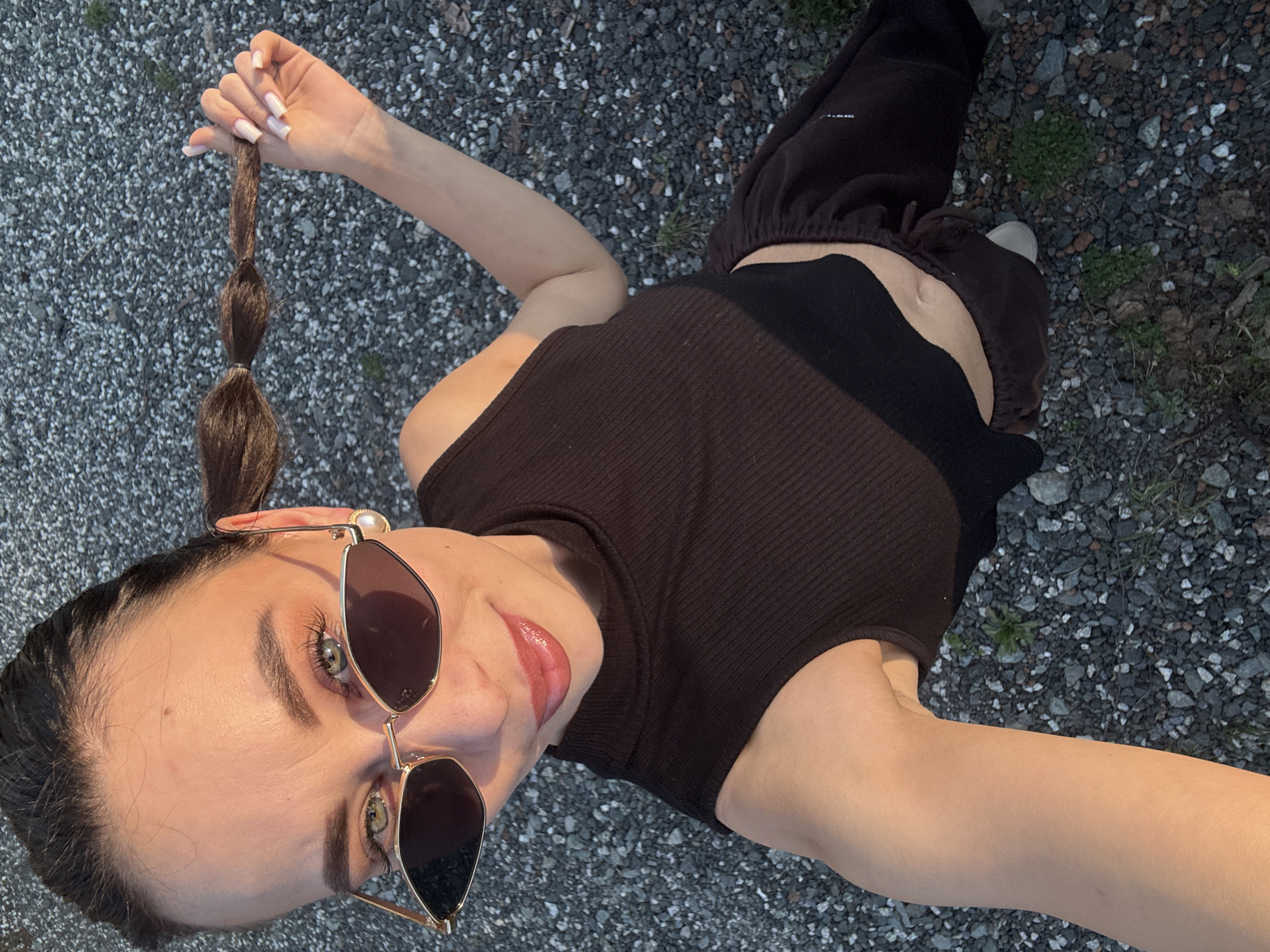 A person with dark hair styled in a bubble ponytail, wearing brown sunglasses, a dark tank top, and dark pants, is lying on a gravel surface. The person is looking up and holding their ponytail. The location appears to be outdoors. The city and country (Chaikovsky, Russia) are mentioned for context, but no elements of the scene obviously reflect or indicate that location.