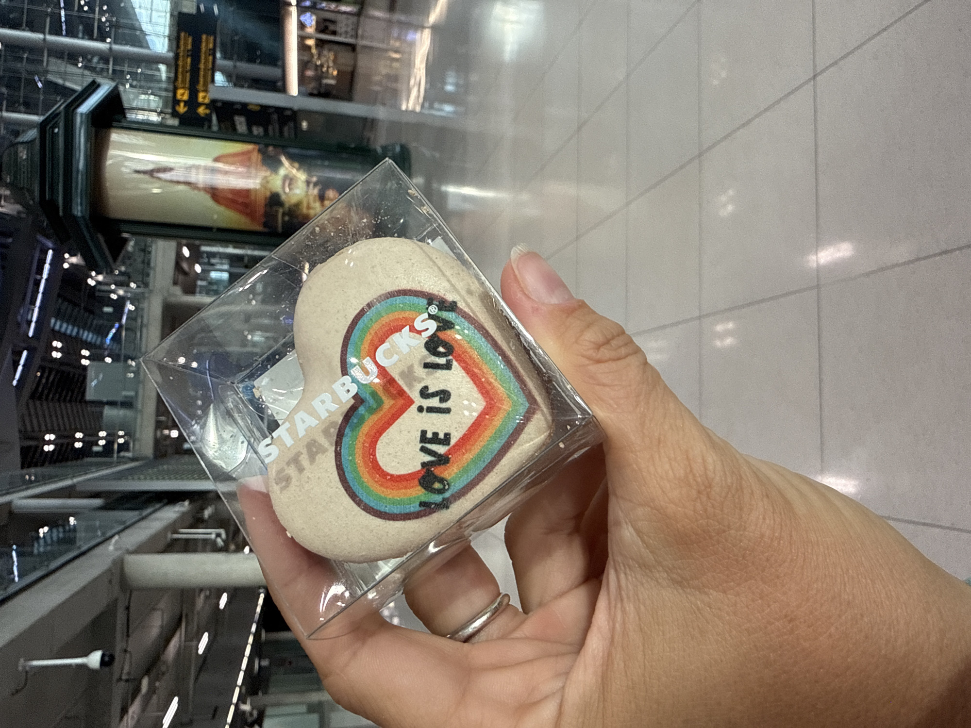 A person's hand, adorned with a silver ring on the index finger, holds a heart-shaped cookie in a clear plastic box. The cookie features a rainbow stripe design with the words "STARBUCKS" and "LOVE is LOVE" printed on it. The background suggests an indoor public space, possibly an airport or large shopping mall, with tiled flooring, architectural elements, and signage visible. One sign features Thai text and arrows, indicating a location within Nong Prue Subdistrict, Thailand.