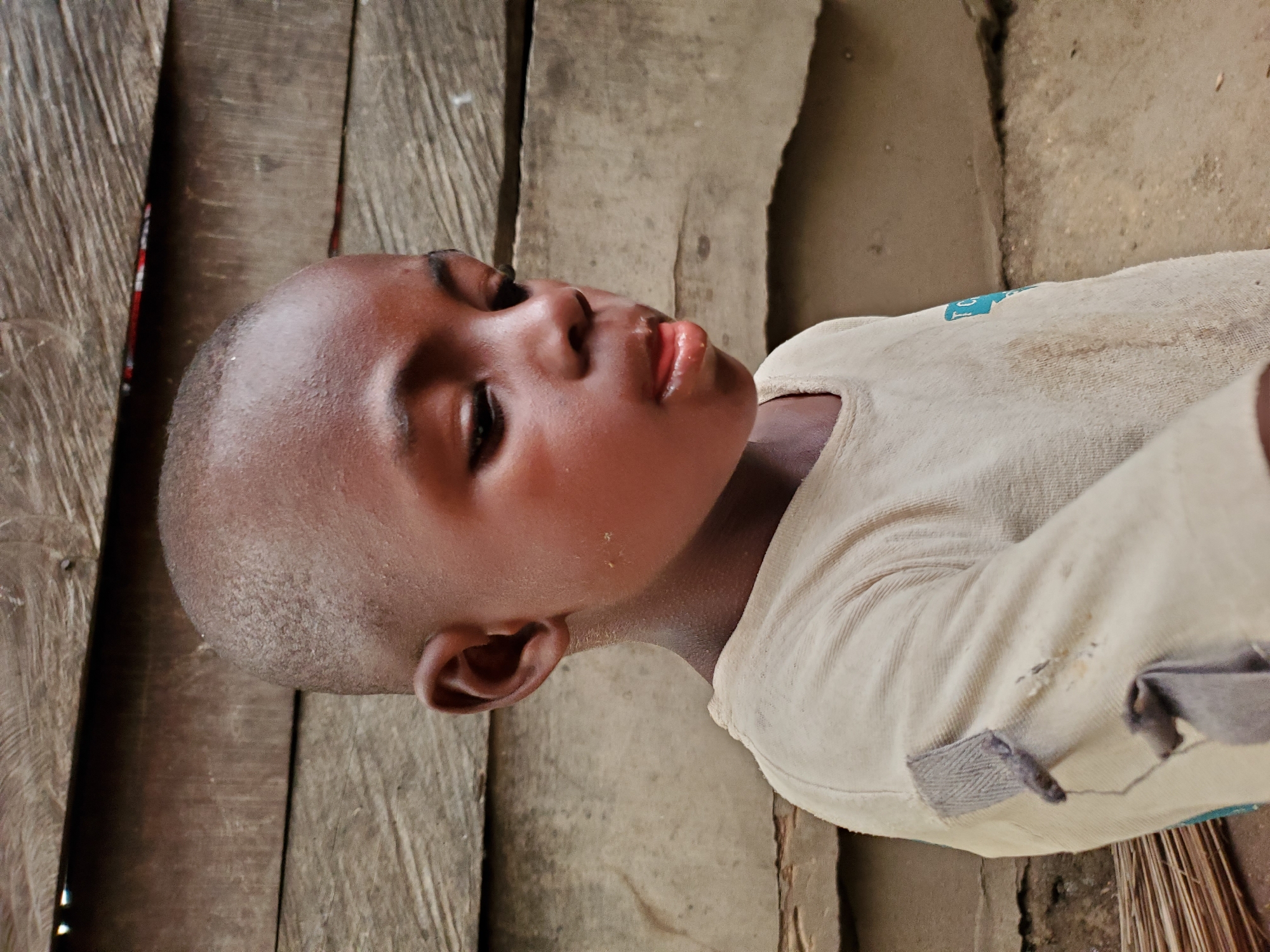 A close-up shot of a young boy is captured indoors. The boy has a shaved head and dark skin. He is wearing a light-colored, long-sleeved shirt. The scene includes wooden planks, forming a wall or floor, along with a dirt or concrete surface. The image suggests a simple, perhaps impoverished, environment. While the specific location in Mararaba, Nigeria cannot be determined from the image, the boy's appearance and the surroundings are consistent with the region.