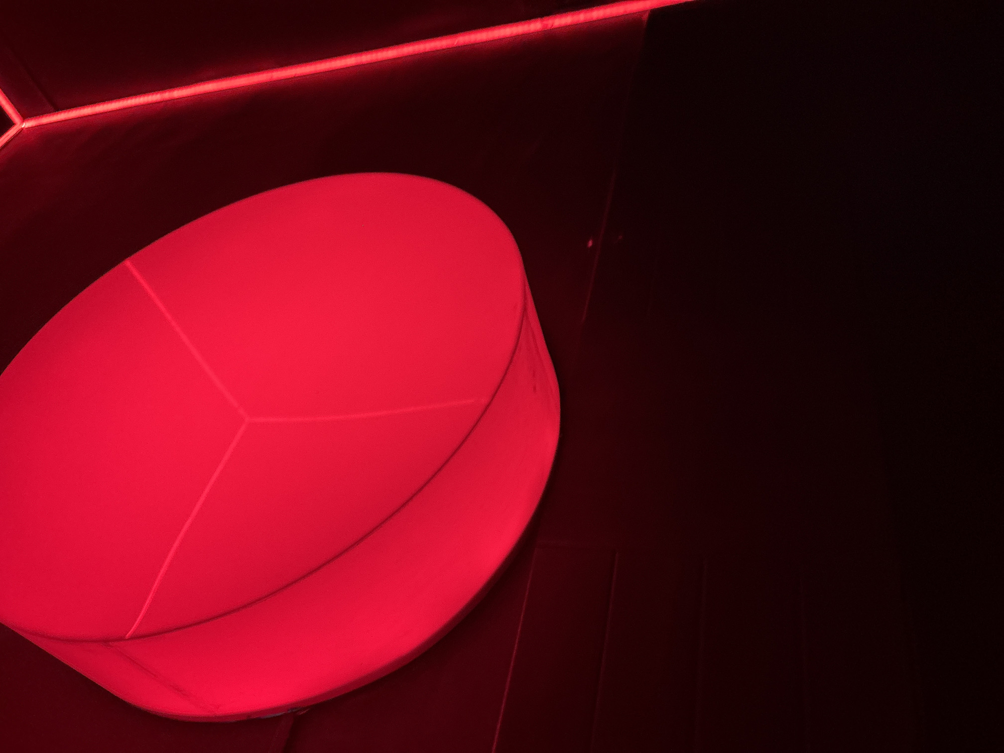 An overhead shot shows a large, circular, red-lit fixture against a dark background. The fixture is illuminated from within, casting a vibrant red glow that highlights its curved surface and an internal geometric design. To the right of the fixture, a narrow, vertical strip of red light is visible, suggesting an accent light or architectural detail. The overall scene is dark, with the red light being the dominant source of illumination, creating a dramatic and modern aesthetic. The location is identified as North Kuta, Indonesia.