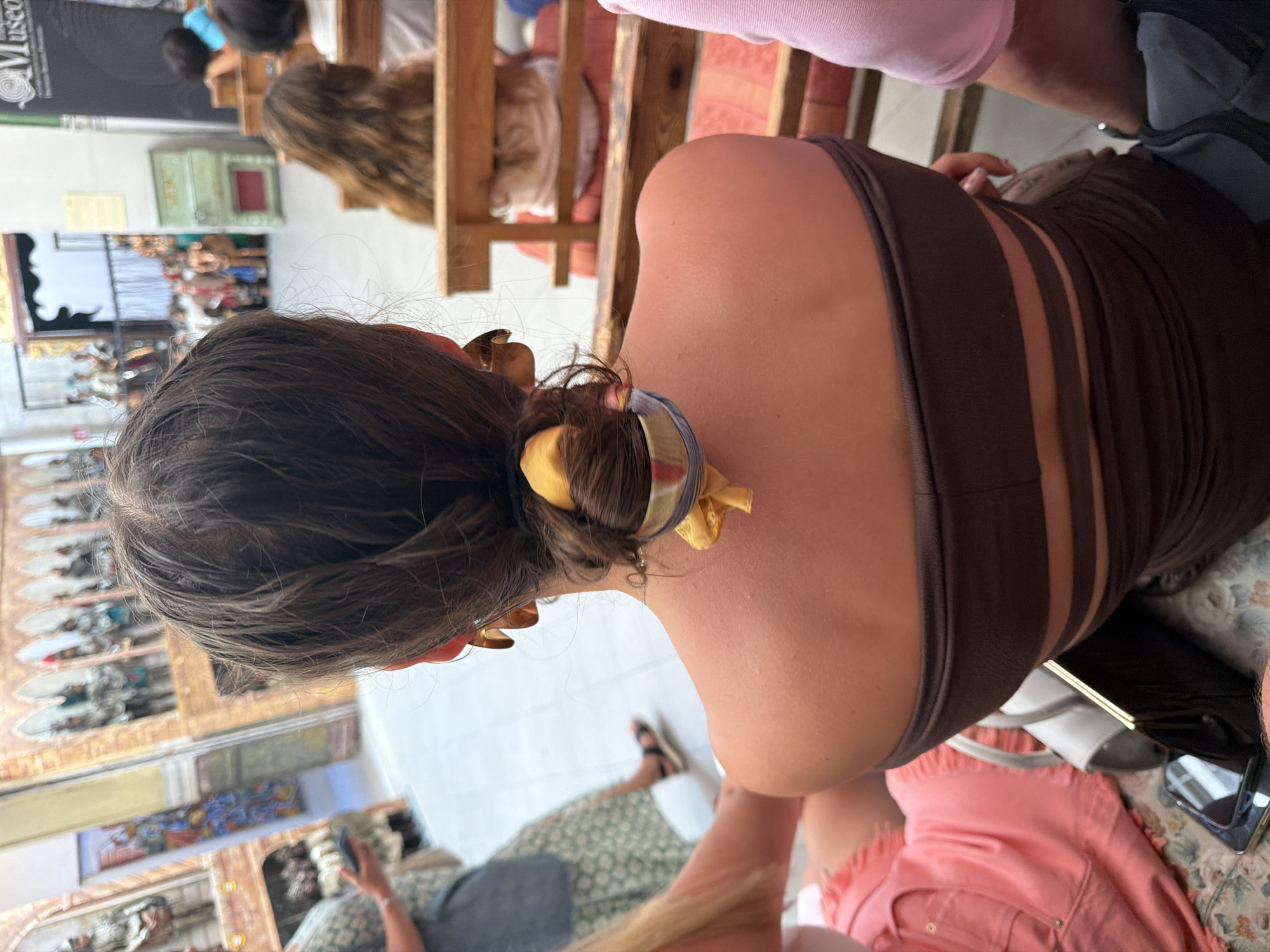 The image captures a rear view of a young woman with dark hair styled in a bun, adorned with a yellow scarf and large gold earrings. She is wearing a brown strapless dress with cut-out detailing. To her left, another woman in a pink top and a person in a light-colored dress are visible, as well as a person in a light pink garment to the right. The setting appears to be a museum or exhibition space, with displays of mannequins in historical attire and architectural elements reminiscent of arches in the background. The floor is tiled, and wooden benches are present. The overall atmosphere is casual, suggesting a leisurely outing. The exact location in Palermo, Italy, is not identifiable from the visual information provided in the image.