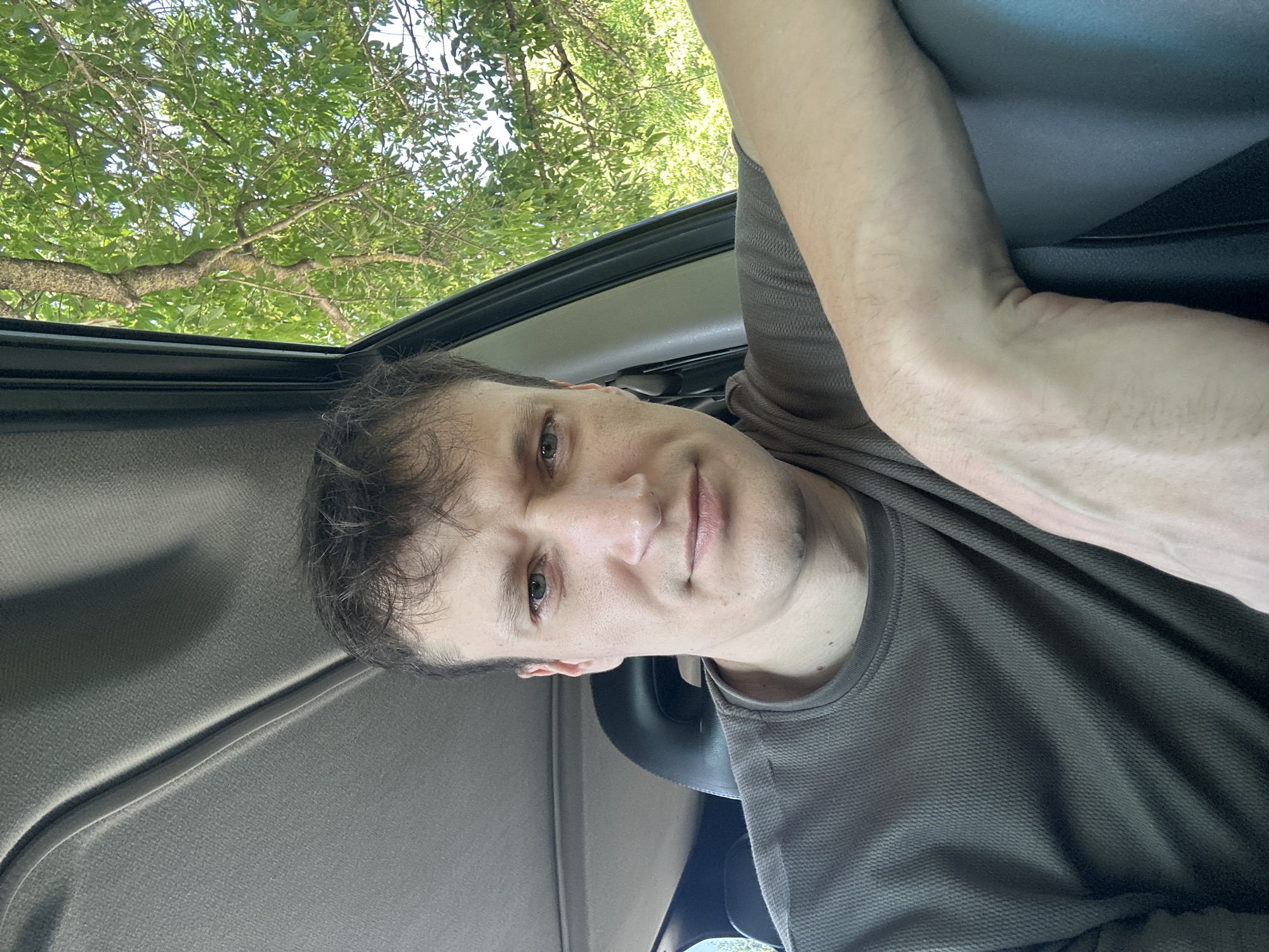 The image shows a medium shot of a man sitting in a car, looking directly at the camera. He has short, dark hair, fair skin, and light-colored eyes. He is wearing a dark gray, textured crew-neck t-shirt. His right arm is resting on the door panel of the car. The interior of the car, including the headrest and ceiling, is visible. Through the open car window on the right side of the frame, lush green trees and foliage are visible, suggesting an outdoor setting with abundant vegetation. The lighting indicates it is daytime. The scene appears to be in Makiv, Ukraine, though no specific landmarks are identifiable in the provided image.