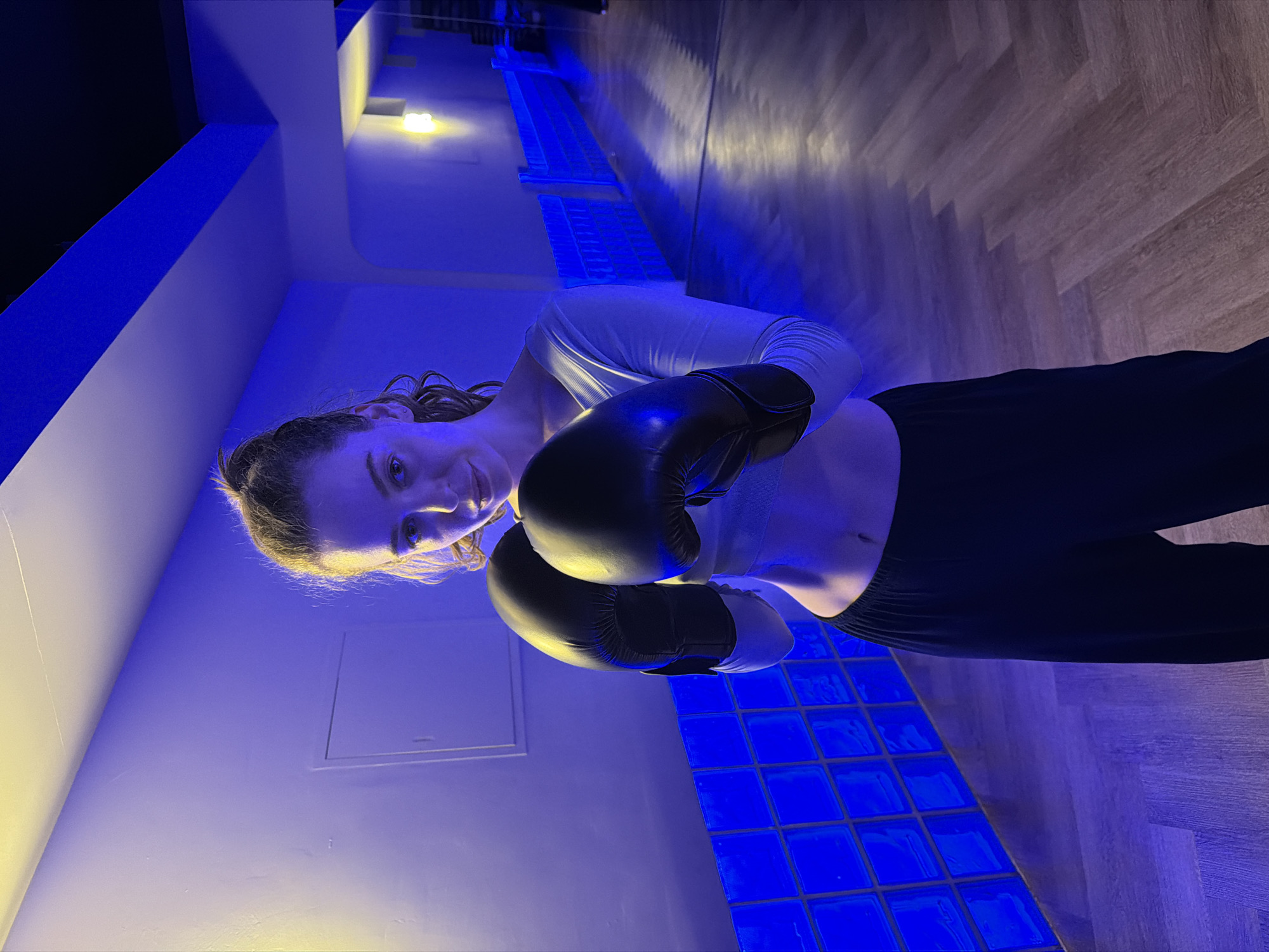 A young woman wearing a light-colored long-sleeved shirt and black pants is wearing black boxing gloves and smiling. She is standing indoors with blue and yellow lighting. A wall with a row of lights is in the background. The floor has a blue grid pattern. The location is Moscow, Russia.