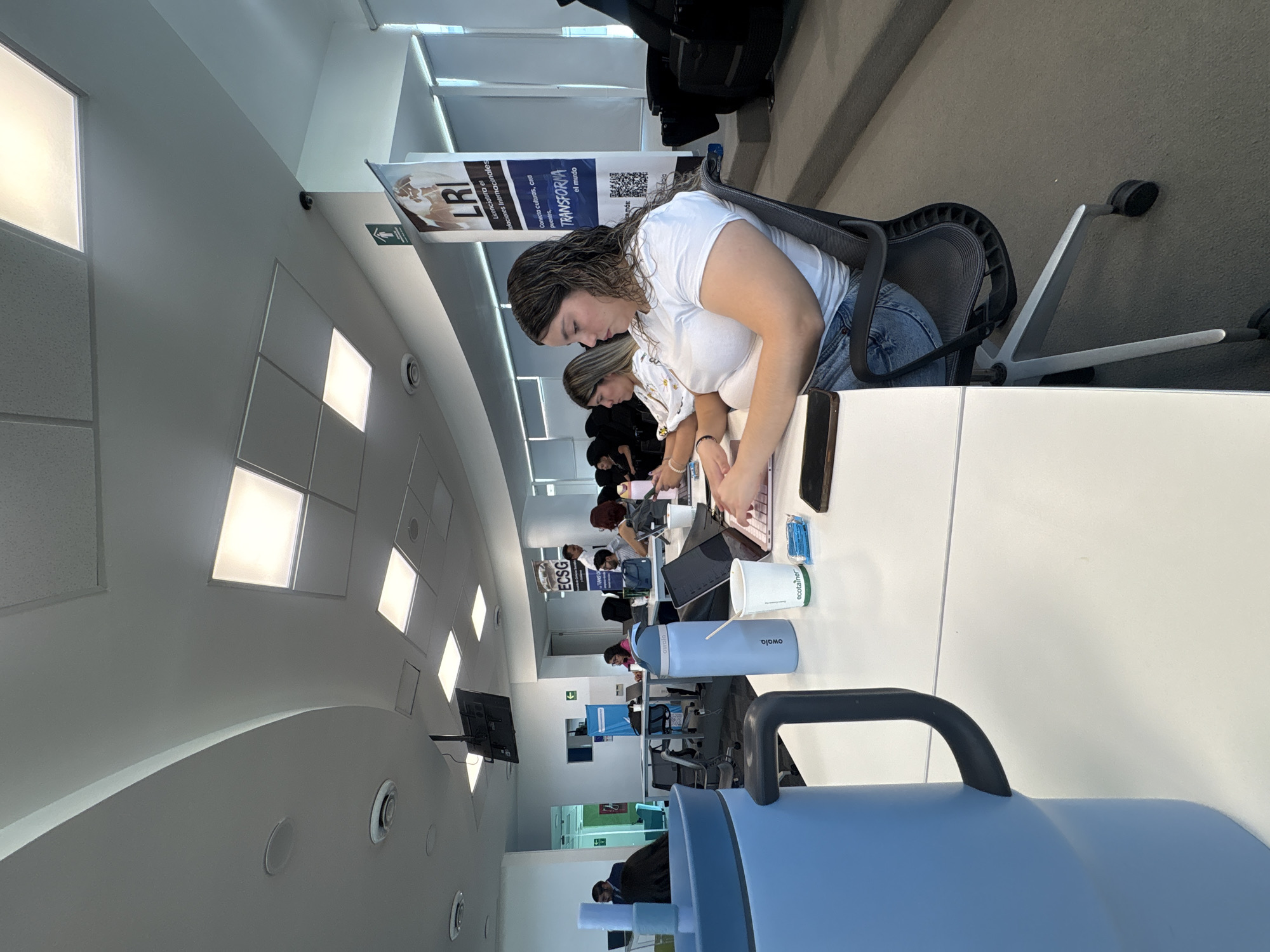 The image shows a group of people sitting at tables in what appears to be a classroom or conference room. The room has bright fluorescent lighting and a curved ceiling. Several individuals are focused on laptops or other materials on the tables. In the foreground, a large blue water bottle dominates the view. The tables are white and clean, with various items like coffee cups, phones, and papers scattered on them. Banners or posters are visible in the background. The room appears to be modern and well-lit. The location is potentially in the city of Puebla, Mexico, given the signs in the background.