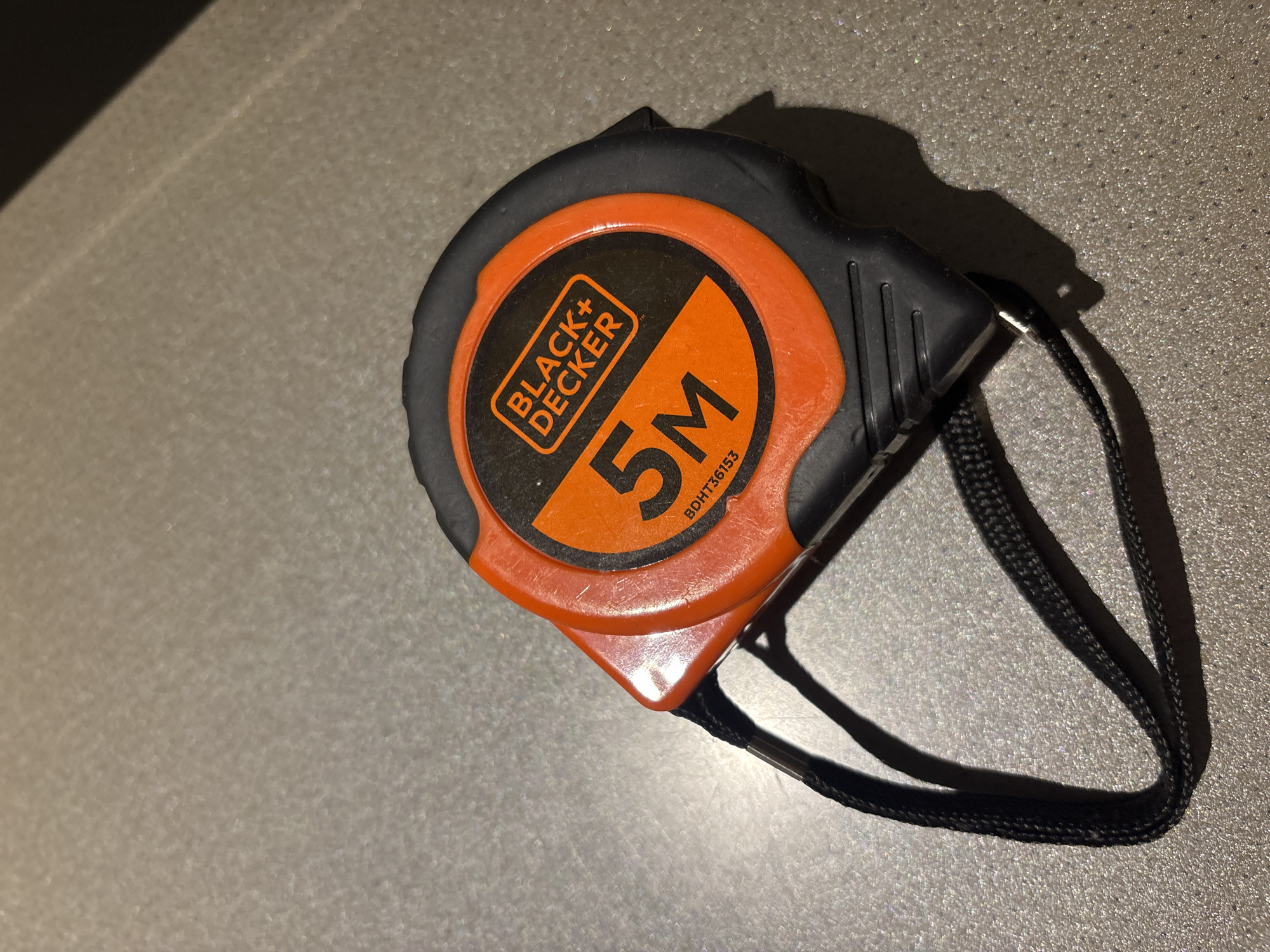 A Black+Decker 5-meter tape measure is resting on a textured, grey surface, possibly a countertop or table. The tape measure is predominantly orange and black. The Black+Decker logo is visible in white lettering on an orange background. The number "5M" is prominently displayed in large, bold black numerals on an orange background. The tape measure has a black lanyard attached. The lighting suggests an indoor setting, with a shadow cast to the right of the object. The surface appears to be speckled, with small reflective particles. There are no people or any discernible Dubai landmarks present in the image.