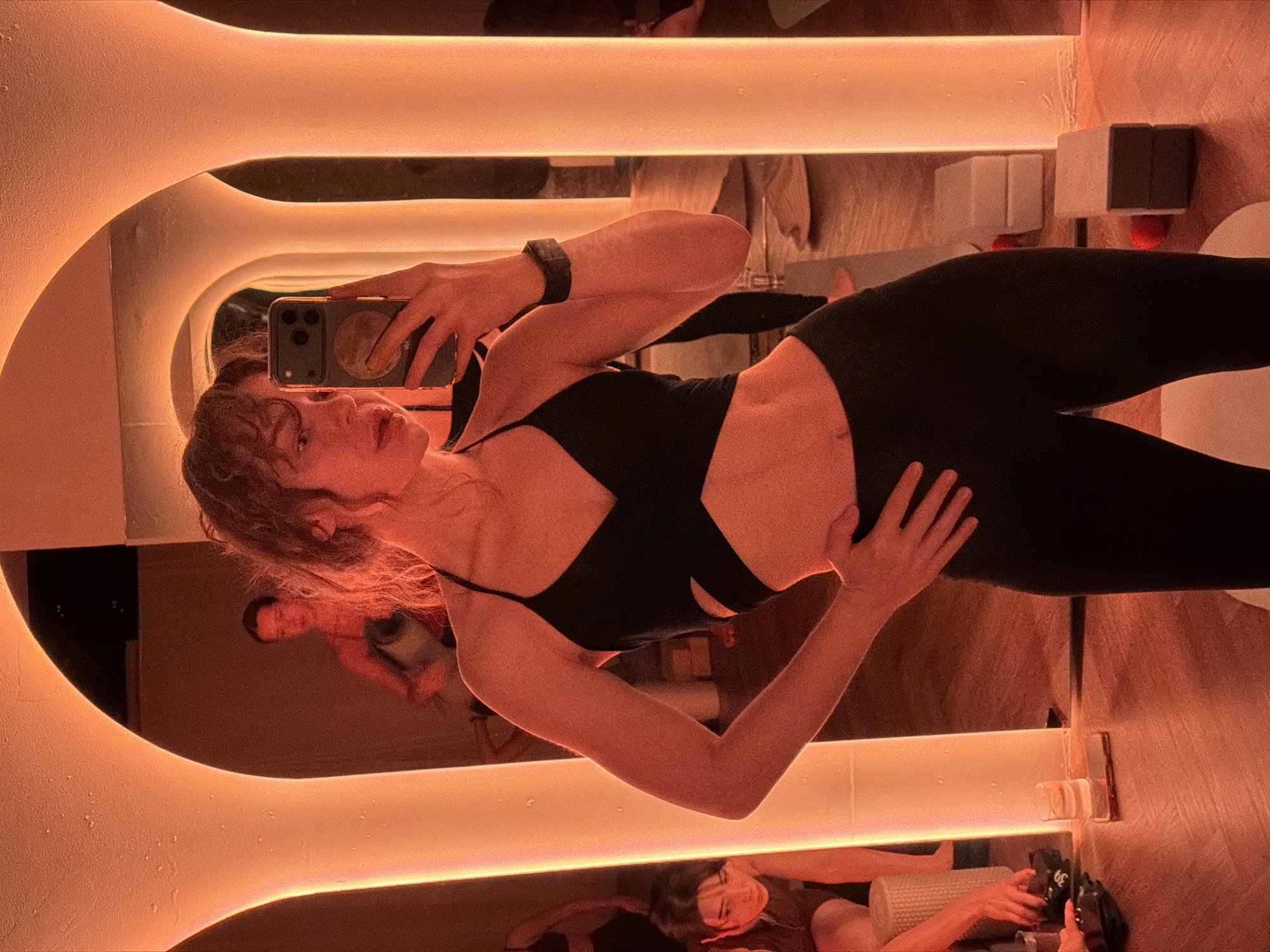 The image captures a young woman taking a mirror selfie in a fitness studio in Moscow, Russia. She is shown from the waist up, holding a smartphone with a circular design on its case to capture her reflection. She wears a black athletic top with a distinctive cross-strap design, black leggings, and a smartwatch on her left wrist. Her dark, wavy hair frames her face as she looks into the phone.
The setting is an indoor gym or studio featuring a large mirror with a unique, U-shaped or wavy illuminated frame that casts a warm, orange-pink glow across the scene. This ambient lighting creates an atmospheric and modern feel, suggesting an evening or night-time workout. The surrounding walls and surfaces also appear to be warm-toned.
In the reflections within the mirror, other individuals are partially visible in the background, including one person with dark hair positioned lower in the frame. Various pieces of gym equipment are also reflected, such as a white foam roller and a black item that clearly displays the number "30". The woman's posture is confident, and her expression is focused on taking the photo.