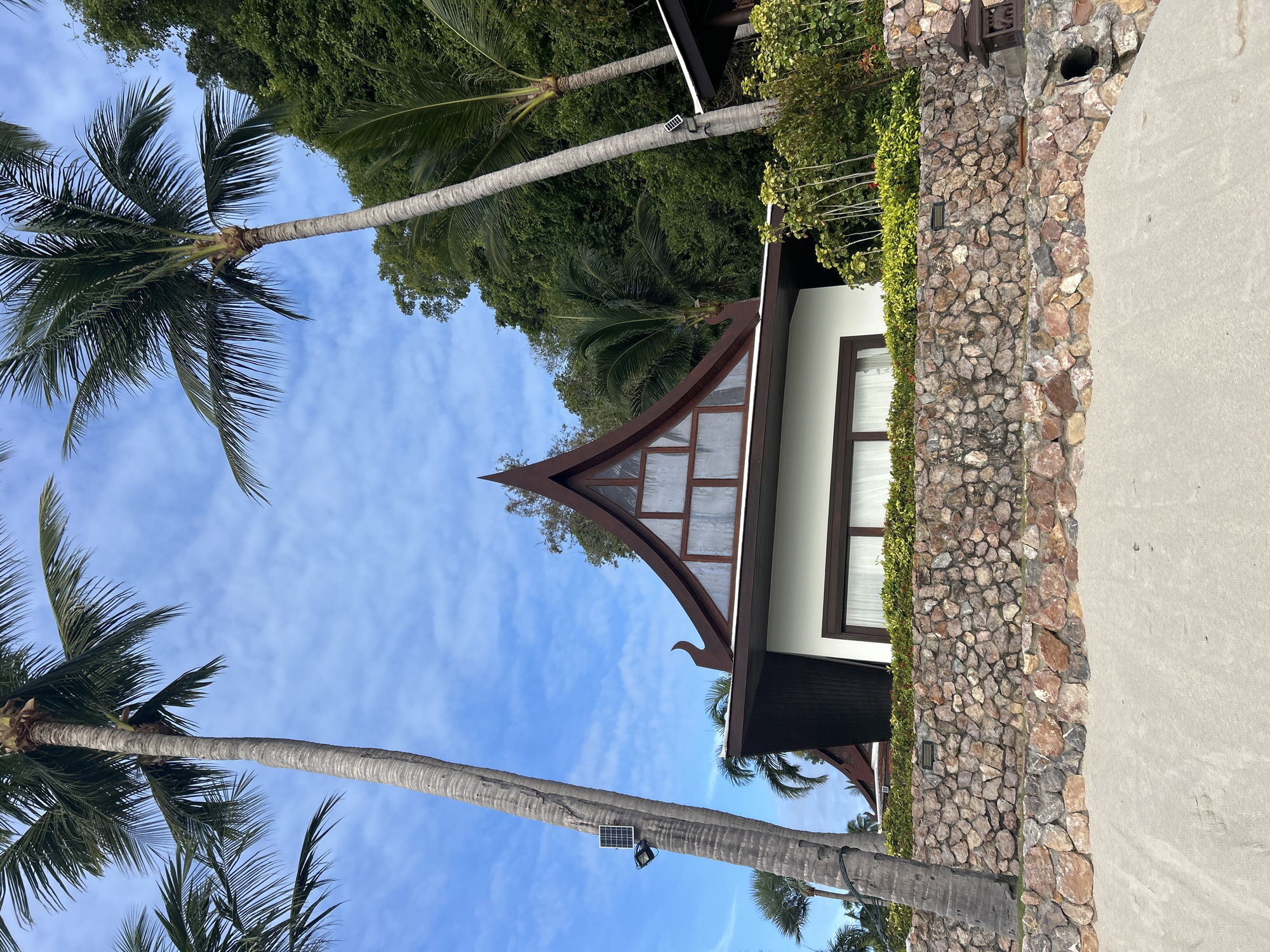 In Ko Samui District, Thailand, a tropical resort building with a distinctive tiered, dark brown roof is visible. The structure is supported by a rough-hewn stone wall that fronts a sandy beach. Two tall palm trees flank the building, their fronds reaching towards a partly cloudy blue sky. A solar panel and a light fixture are attached to the trunk of the left palm tree. The building has white walls and dark brown window frames. Lush green vegetation covers the hillside behind the resort.