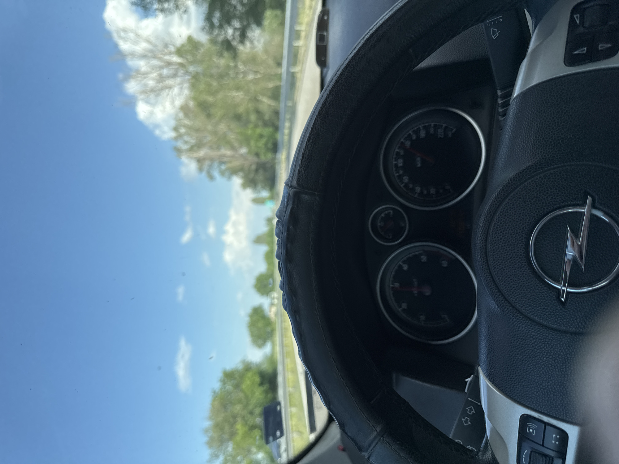 The image shows the interior of a vehicle, focusing on the steering wheel bearing an Opel logo and the dashboard with speedometer and other gauges. The view through the windshield reveals a daytime scene of a road lined with trees under a blue sky with scattered clouds. The location, Kalugerovo, Bulgaria, is based on a search of the logo on the steering wheel. No people are visible. The car appears to be in motion.