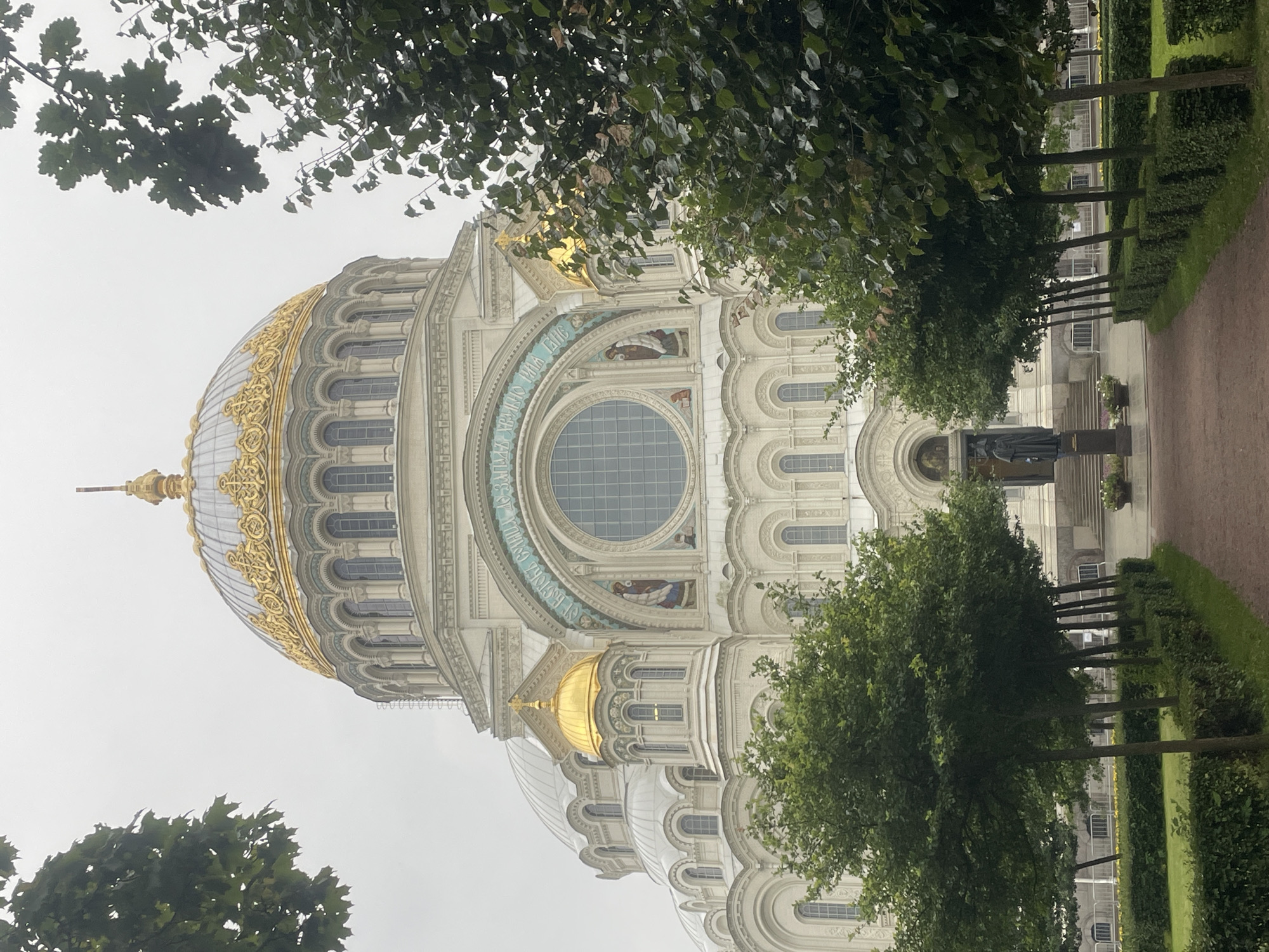 The image shows the Kronstadt Naval Cathedral, a large Orthodox cathedral located in Kronstadt, Russia. The cathedral is white with a large, golden dome. It is surrounded by trees and manicured hedges. In front of the cathedral is a statue of a man in a dark robe. The sky is overcast and the overall lighting is subdued.