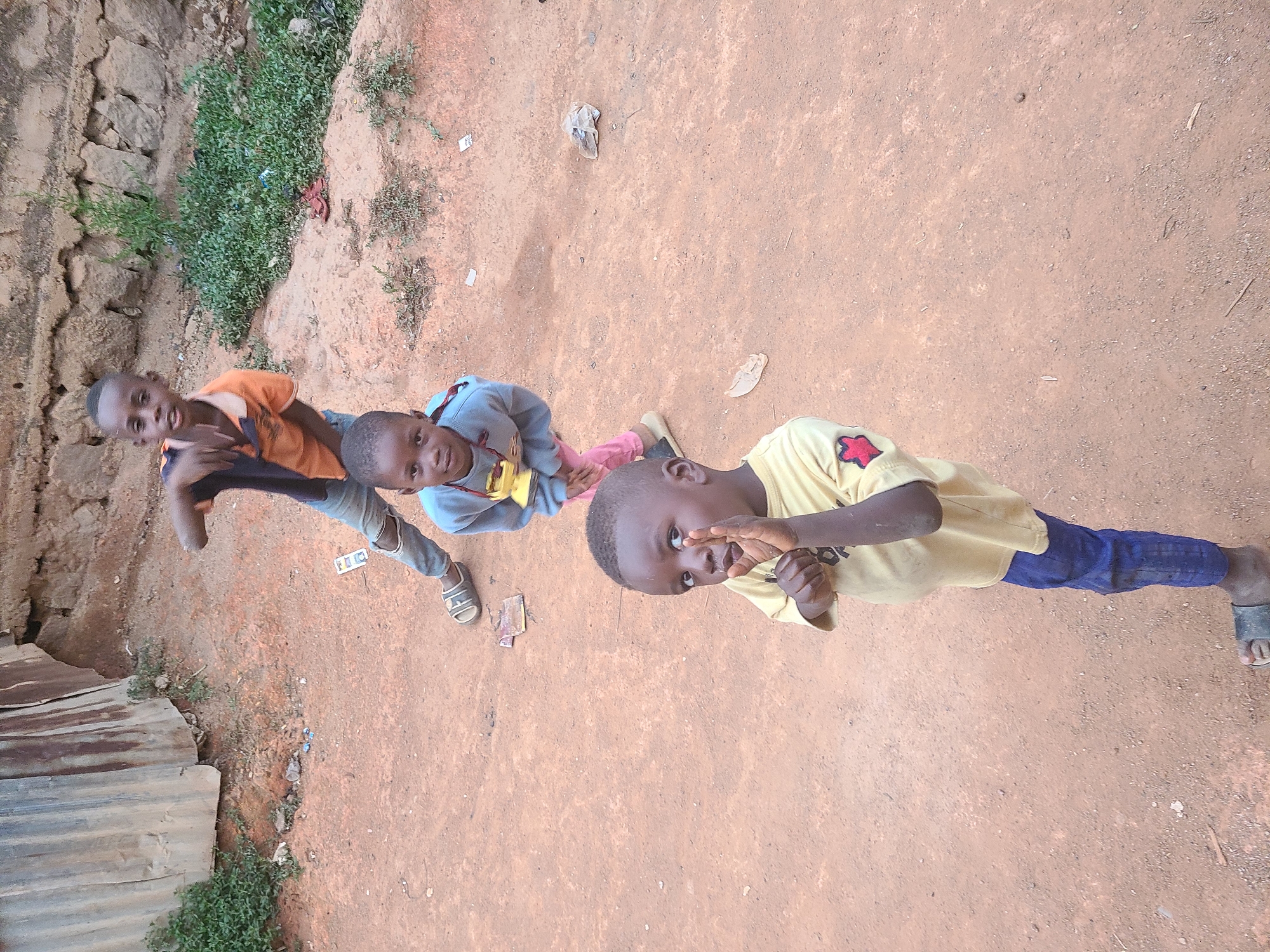 Three Black children are standing in an unpaved area in Jos, Nigeria. The child closest to the viewer wears a yellow shirt and blue jeans. The middle child wears a blue top and pink pants. The child furthest away wears an orange top and grey pants. All three children are looking up towards the camera. There is a wall on the left side of the image. There is vegetation growing on top of the wall and along the side of the unpaved area. There is corrugated metal in the bottom left corner. There is loose trash on the ground. The unpaved area is red-brown dirt.