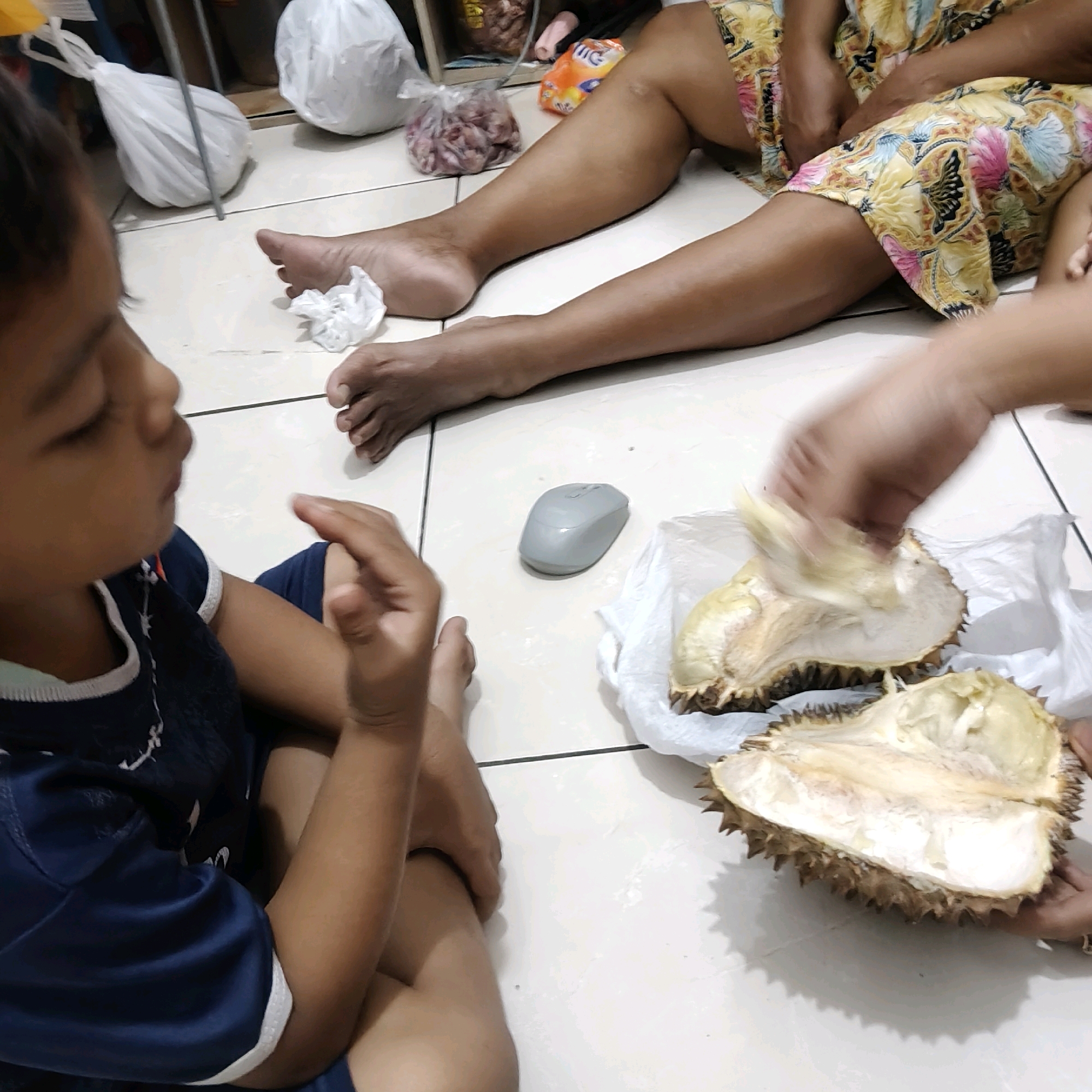 A child in a dark blue shirt sits on a tiled floor, facing left. A person's legs, wearing a colorful patterned fabric, are visible on the right side of the frame. In front of the child, two halves of a durian fruit are open, resting on white paper. A gray computer mouse is located to the left of the durian. In the background, white plastic bags and other items are stored. This scene appears to be indoors, possibly in a home in Mekarmukti, Indonesia, with the floor being made of light-colored tiles.