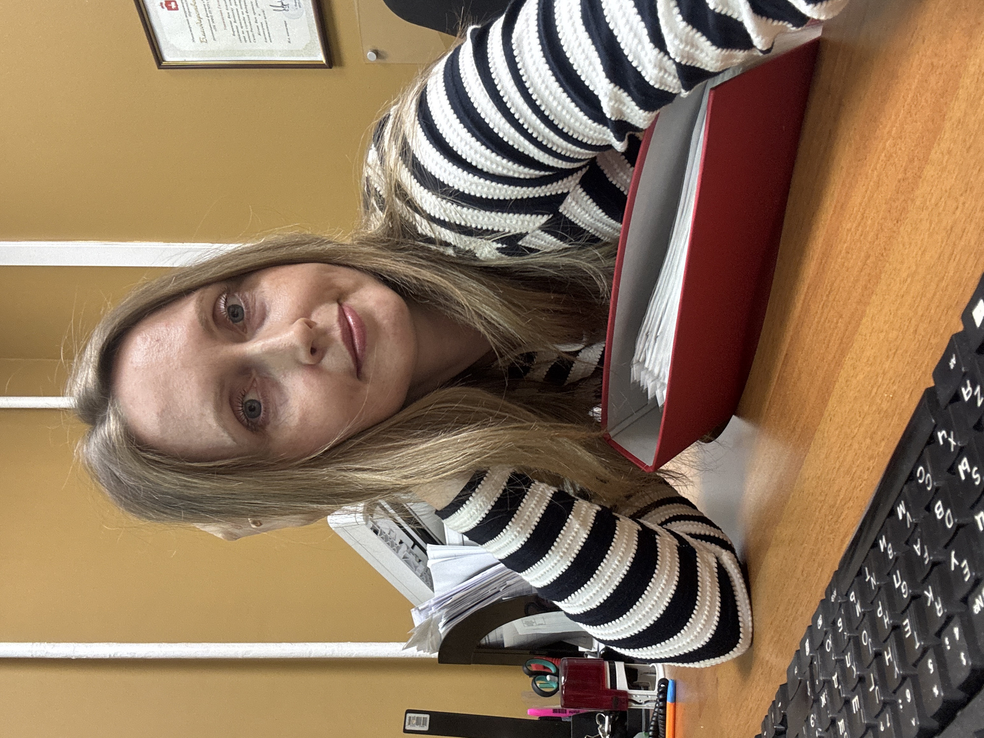 A woman with long blonde hair is seated at a desk. She is wearing a black and white striped sweater. A red file box filled with white papers is on the desk next to her, along with a black computer keyboard. In the background, a document in a frame is mounted on a beige wall. There is also a printer, writing supplies, and an in tray. The specific location in Chaikovsky, Russia cannot be determined from the image.