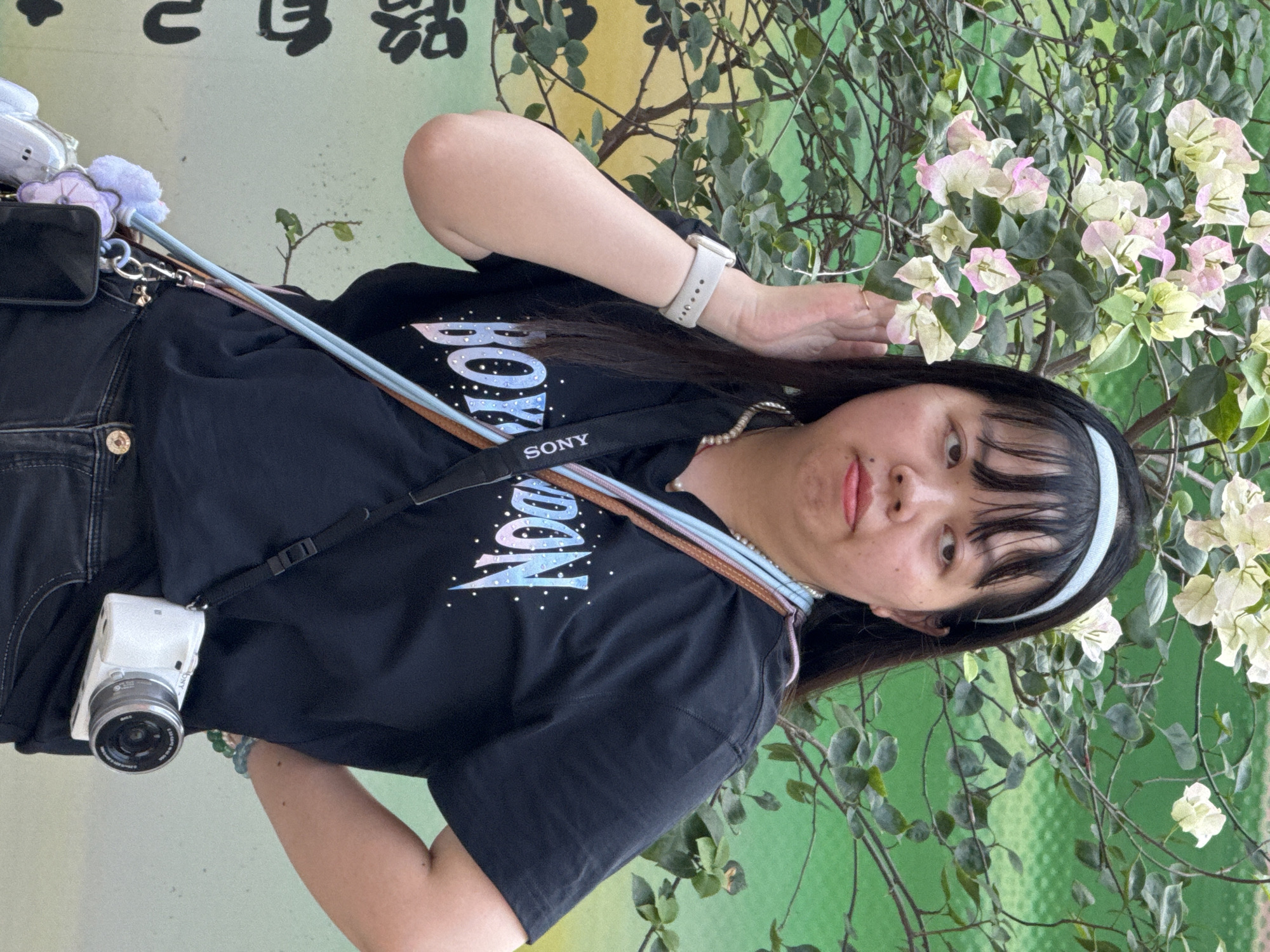 An East Asian woman is captured in a portrait, likely outdoors in Yuzhong District, China. The image is rotated counter-clockwise, showing her with her head tilted, looking directly at the viewer with a slight pout. She has dark hair with bangs, held back by a light blue headband. She wears a black t-shirt featuring a colorful, gradient graphic that partially reads "BOY" with white speckles, and black jeans.
Numerous accessories are visible: a white SONY digital camera hangs from a strap across her body, a white smartwatch is on her left wrist, and a pearl necklace adorns her neck. A black bag and several small, colorful charms or keychains are attached to her waistline. Her left arm is bent, with her hand near her ear/hair, while her right arm is out of frame.
The background consists of a green-yellowish wall partially obscured by abundant green foliage and branches. Clusters of white and light pink flowers, resembling bougainvillea, are interspersed among the leaves. In the upper left corner, dark Chinese characters are visible on the wall. The scene appears to be set during the daytime.