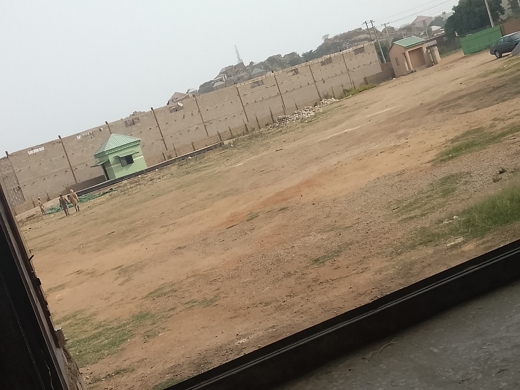 The image captures an expansive, semi-developed outdoor area, likely a construction site or undeveloped land, viewed from behind a dark foreground frame, possibly a window. The scene is dominated by a large, reddish-brown dirt field with patches of sparse green grass.
The field is partially enclosed by a tall, unfinished concrete block wall that stretches across the mid-ground and background. The wall features visible rebar or support posts at regular intervals. Behind this wall, on distant hills, several residential buildings are visible, along with a prominent radio tower or similar mast.
In the mid-ground, to the left, stands a small, light green building with a darker green, pitched roof, resembling a guard post or shed. Further back and to the right, another small, brown building with a green roof is partially visible near the wall. A dark-colored car is parked in the far background, behind a green fence or gate.
Several individuals, at least three, are present in the left-mid ground of the field, near the green structure. They appear to be walking or standing, but their specific activity is not clearly discernible.
The time of day is daylight, characterized by bright but diffused light and a pale, hazy sky, suggesting an overcast day or possibly dusty conditions. The overall atmosphere is utilitarian and somewhat desolate. There is no legible text visible within the image.