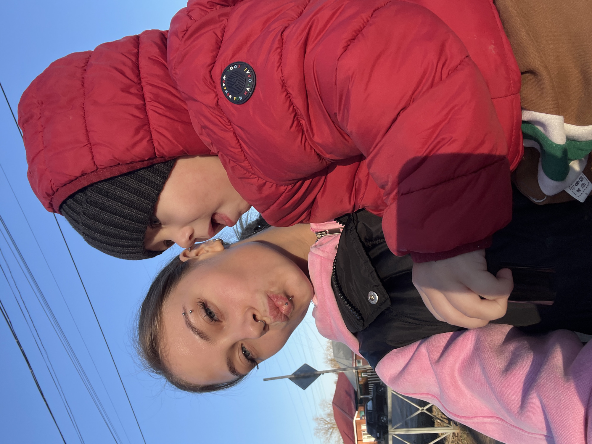 An outdoor, eye-level photograph, rotated 90 degrees clockwise, captures two individuals against a clear blue sky. The scene is set in what appears to be a street or residential area, indicated by distant buildings, power lines, and a partially visible road sign.
The primary subjects are an adult, likely female, and a young child. The adult, positioned in the foreground, has dark hair, blue eyes, and multiple facial piercings including eyebrow, septum, philtrum, and lower lip. She is wearing a pink hooded sweatshirt under a black vest and has a playful expression with puckered lips.
The child, in the background and slightly to the side, wears a dark knit beanie and a bright red puffer jacket with a hood. A brown and green striped garment is visible beneath the jacket. On the left arm of the red jacket, there is a circular black patch with white text that reads "COOL WEATHER FUN MAKERS". The child's hand is seen resting on the adult's midsection, possibly holding on. He looks towards the adult with a soft, engaged expression. A white tag with laundry care symbols and a size indication (possibly "110 CM") is visible on the child's brown garment.
The time of day is daytime, evidenced by the bright sunlight casting shadows and illuminating the scene. The weather appears clear and sunny with a vibrant blue sky overhead. The overall mood of the image is casual and affectionate, suggesting a candid moment shared between the two individuals. The location context is Fokino, Russia.