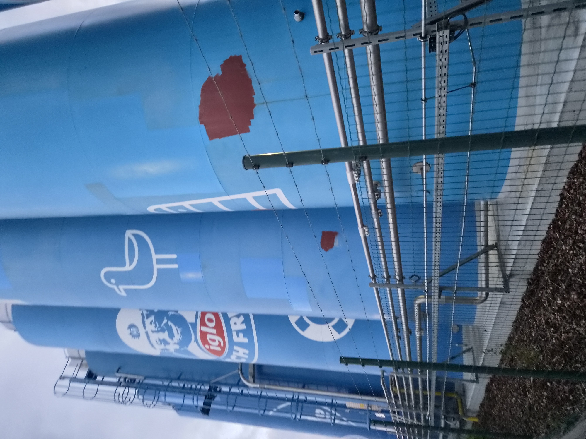The image shows a row of large, blue industrial silos in Bremerhaven, Germany. The silos are cylindrical and appear to be made of metal. They are marked with white logos and symbols. One silo features a cartoonish face with a hat and the text "iglo" and "CH FR". Another silo has a white outline of a seagull. A third silo has a white outline of a life preserver ring. There is a red, irregularly shaped mark on one of the silos.
The silos are situated in an outdoor industrial area. A metal fence with barbed wire runs across the foreground, obscuring a portion of the silos and the ground. The ground in front of the fence is covered with dark mulch and some dead vegetation. Metal pipes and structural supports are visible running along the base of the silos and behind the fence. The sky above is overcast. The overall scene suggests an industrial facility related to food production or storage, possibly for frozen foods given the "iglo" branding, which is a well-known frozen food brand.