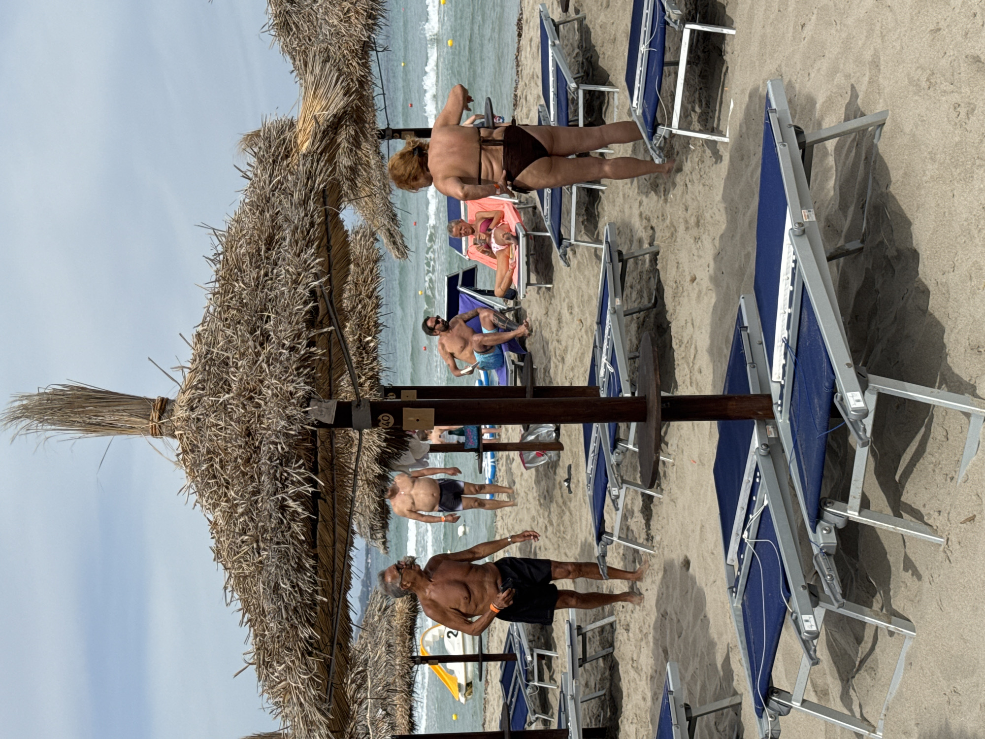 The image depicts a sandy beach scene at Isola delle Femmine, Italy, under a partly cloudy sky. In the foreground, several empty blue and silver lounge chairs are set up on the sand, partially shaded by thatched umbrellas.
To the left, an older man with gray hair and glasses, wearing black shorts, stands near a thatched umbrella, holding an object in his right hand. Behind him, a yellow watercraft with the number "2" on it is visible on the water. Further back, another man, also wearing black shorts, stands near the water.
In the middle ground, a man with sunglasses and a blue towel around his waist sits on a blue lounge chair. Next to him, a woman with blonde hair, wearing a pink bikini top and white bottoms, reclines on a pink lounge chair, looking at her phone. To the right, a woman with reddish-brown hair, wearing a brown bikini, stands facing away from the camera, with her arms slightly bent.
The background shows the blue sea with gentle waves and yellow buoys. The overall atmosphere is that of a relaxed beach resort.