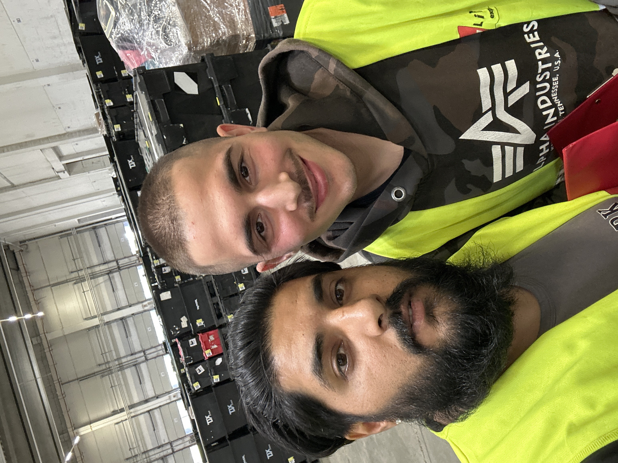 Two men are taking a selfie in a warehouse environment in Schweitenkirchen, Germany. The man on the left has dark hair and a beard, and is wearing a bright yellow vest over a gray t-shirt. The man on the right has short hair and a mustache, and is wearing a camouflage hoodie under a bright yellow vest. They are standing in front of rows of black plastic storage bins, some of which have red accents. The warehouse ceiling is visible in the background, with rows of lights. The overall scene suggests an industrial or logistics setting.
