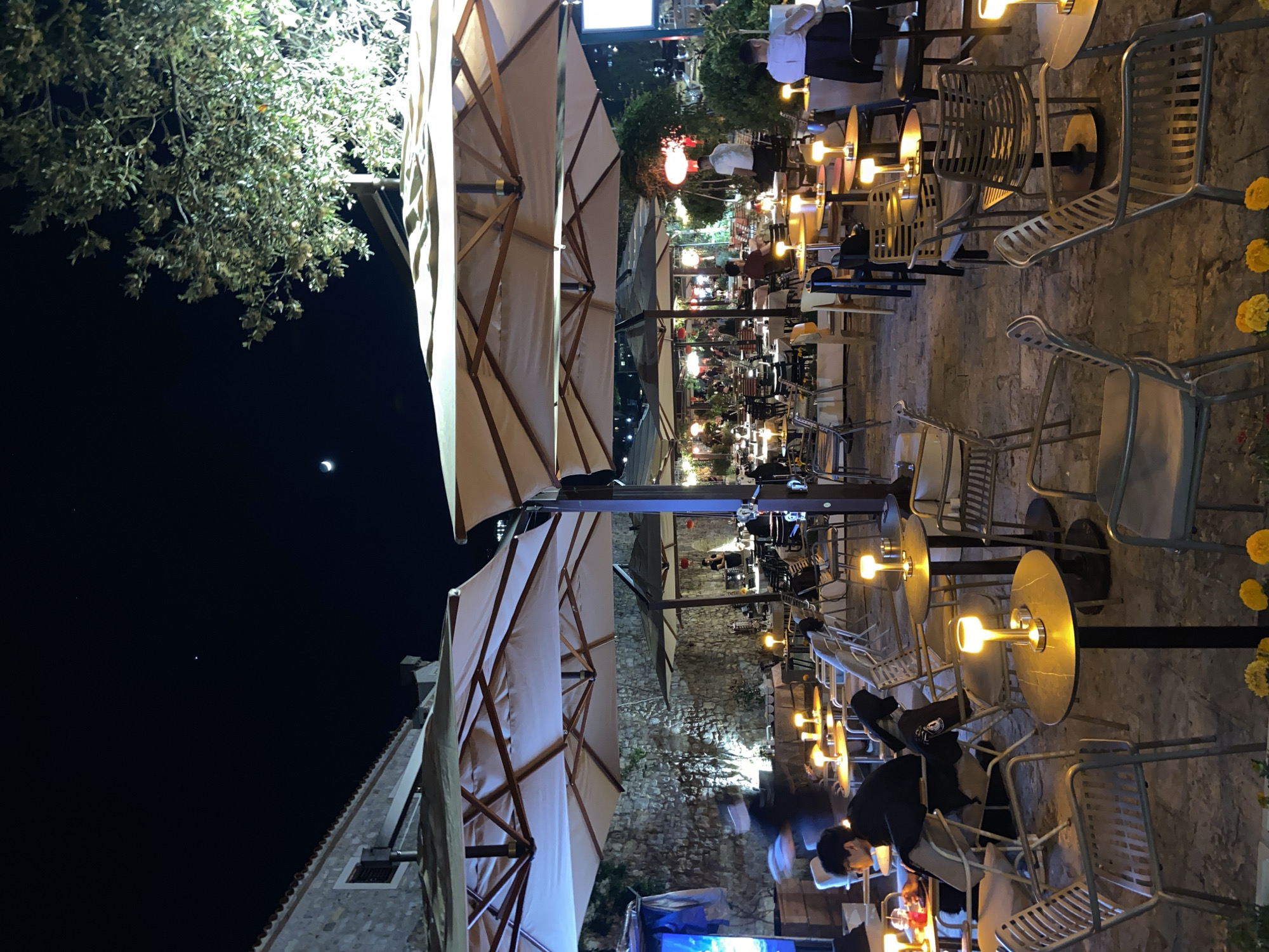 The image depicts an outdoor café scene at night in Budva, Montenegro. Multiple tables with metal chairs are visible, illuminated by table lamps. The tables are arranged along a stone-paved area. Overhead are large, open umbrellas providing shade. Several people are seated at the tables, engaged in conversations or activities like dining. Background features stone buildings and walls, indicative of Budva's Old Town architecture. A crescent moon is visible in the night sky above a tree on the left.