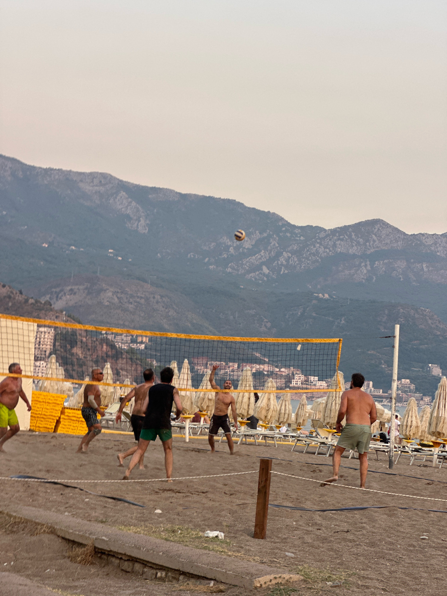 In Boreti, Montenegro, a group of men are playing beach volleyball on a sandy court. The court is bordered by a net with yellow trim and poles. In the background, a line of beach umbrellas and lounge chairs are set up on the sand, with buildings and the Adriatic Sea visible beyond. Further in the distance, rugged mountains rise against a hazy sky. One player is mid-air, about to hit the ball, while others are positioned on the court. The scene depicts a recreational activity in a coastal environment.