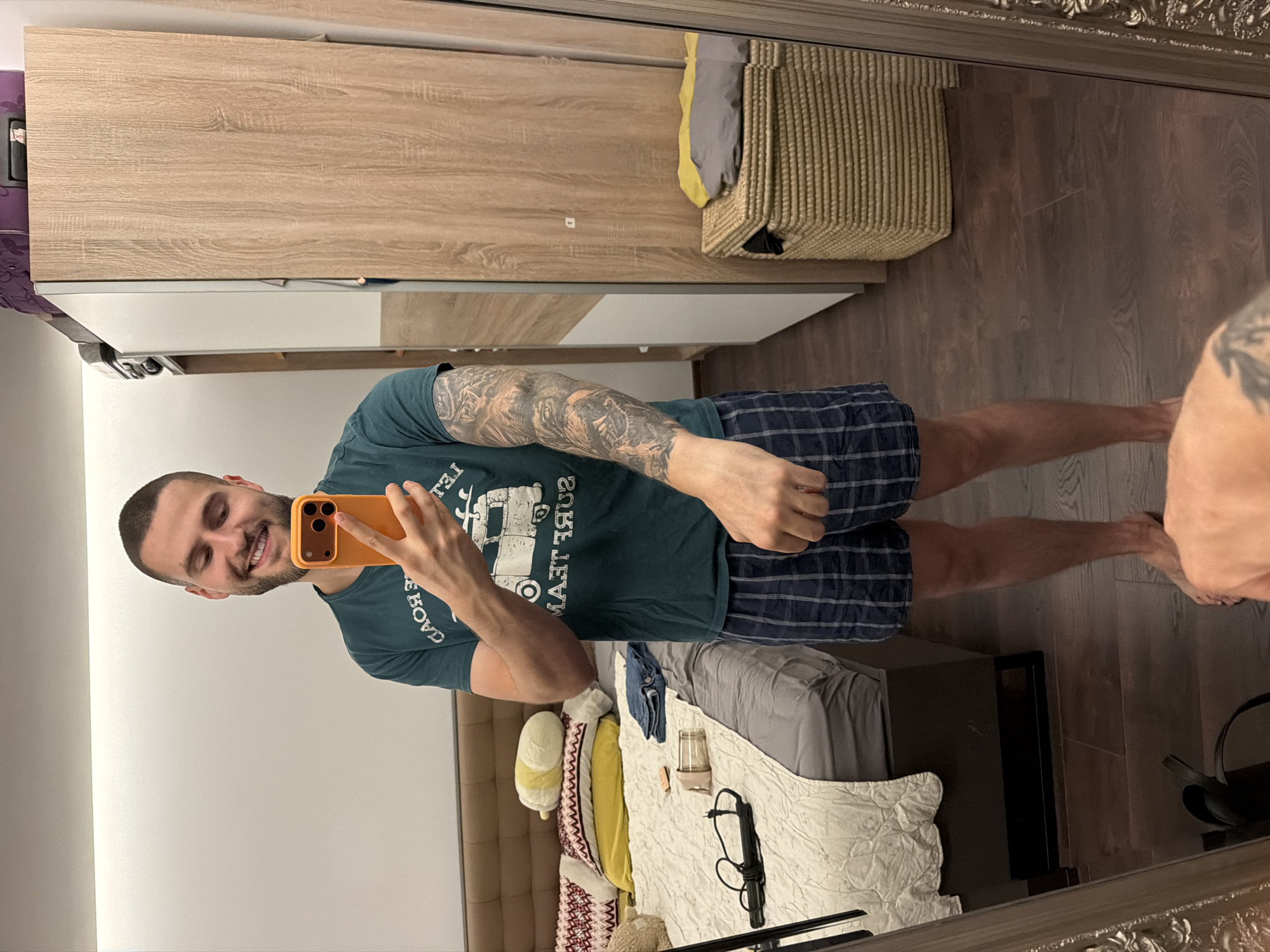 A man is taking a mirror selfie in a bedroom. He is smiling and holding an orange smartphone. He is wearing a dark green t-shirt with a graphic print and blue and white checkered boxer shorts. The bedroom has a bed with pillows, a bedside table with items on it, and a large wardrobe with wooden doors. The floor is dark wood. A woven basket with items on top of it is visible to the right of the wardrobe. The mirror is ornate and framed. The image is captured from the chest down. The man's left arm has a large tattoo. There is no specific indication of the location being in Sofia, Bulgaria.