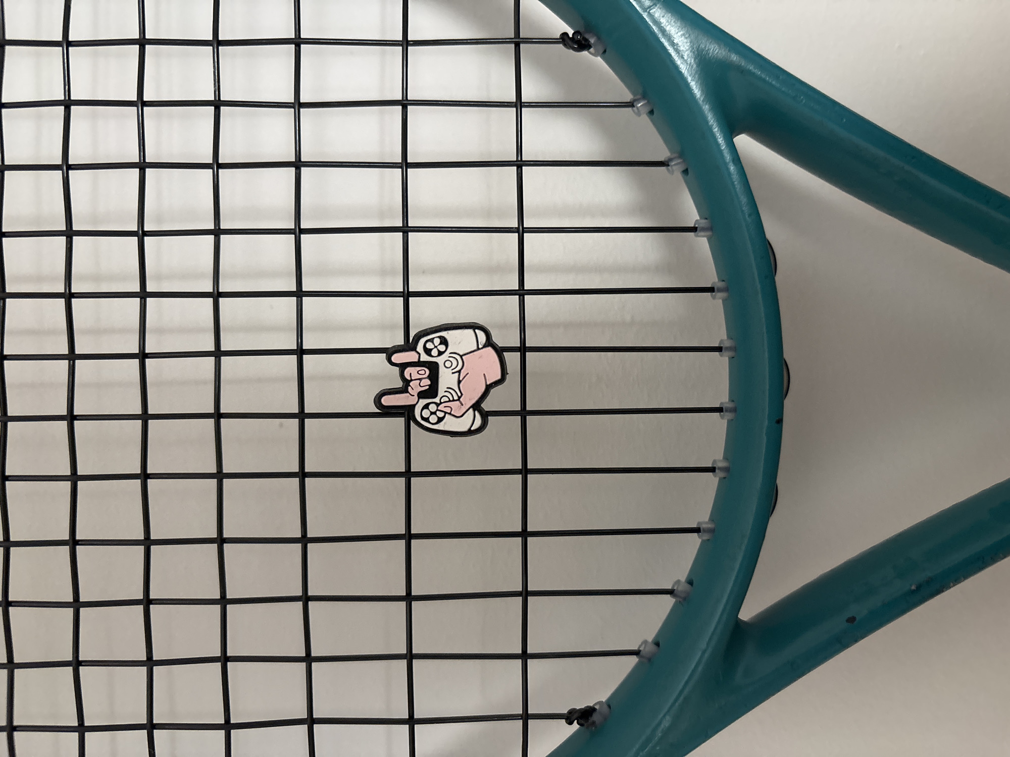 A close-up shot shows a tennis racket with black strings. A cartoon pin featuring a hand holding a video game controller is attached to the strings. The racket frame is a teal color. The background is a plain white wall. The photograph appears to be taken indoors. There is no indication of the city of Chaikovsky, Russia in the image.