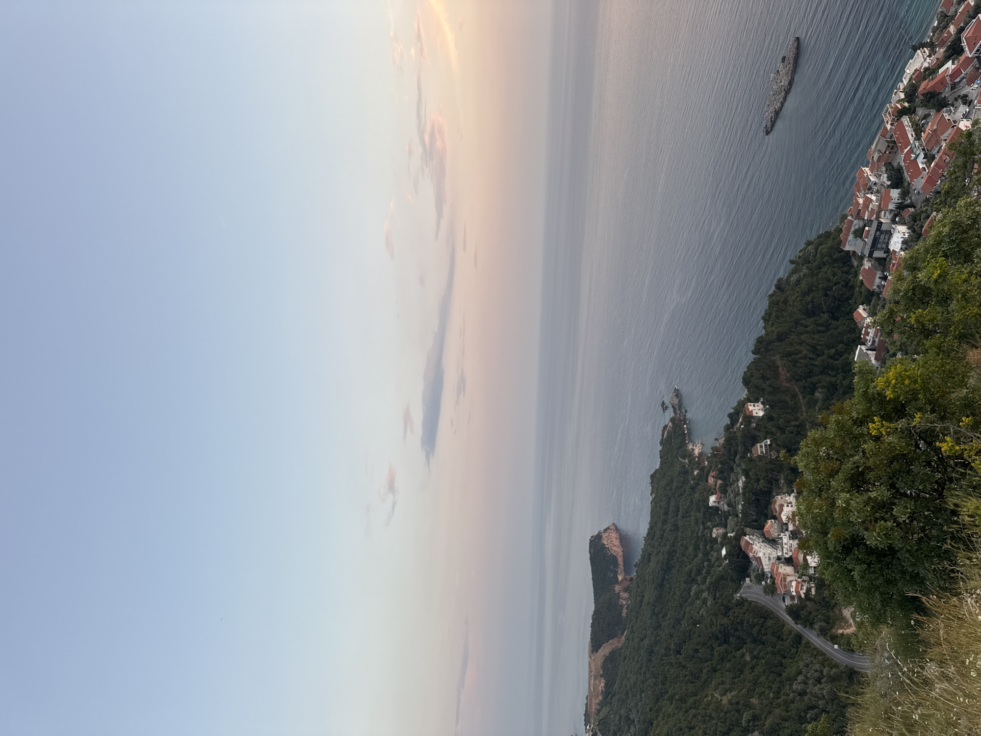 The image depicts a coastal view in Sveti Stefan, Montenegro, likely taken from an elevated vantage point. The sky transitions from a light blue to a pale pink hue, suggesting either sunrise or sunset. The sea occupies a significant portion of the frame, its surface reflecting the sky's colors. Along the coastline, there are clusters of buildings with red-tiled roofs, typical of Mediterranean architecture. Dense vegetation, including trees and shrubs, covers the land near the coast. A road winds its way through the landscape. In the sea, there is a rocky islet near the coastline, and further out, a larger rocky outcrop.