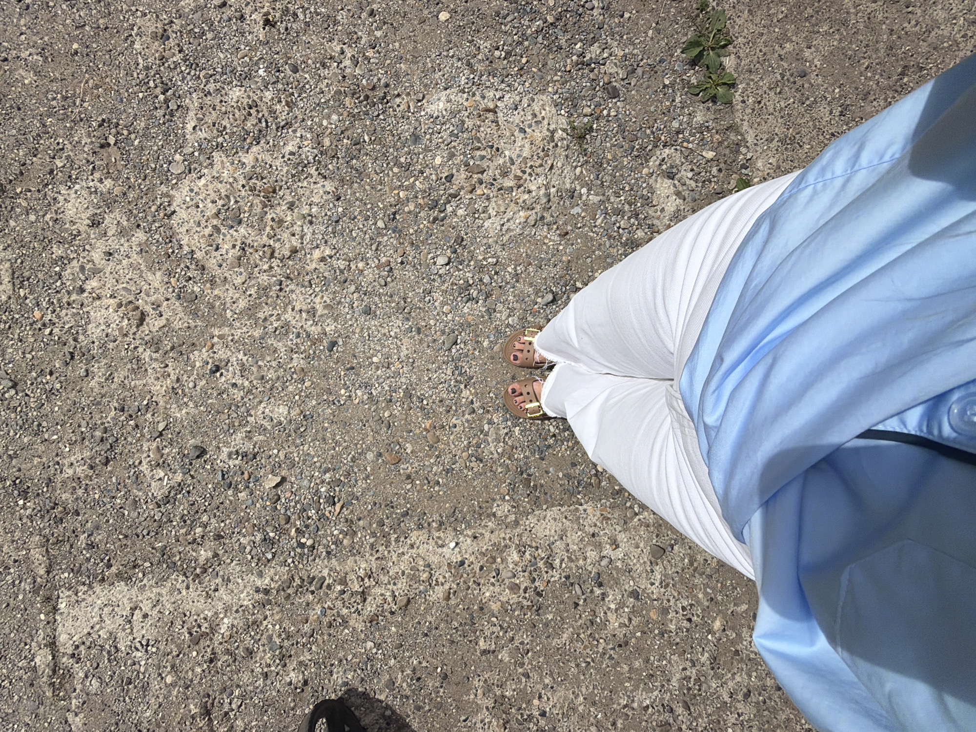 This media file captures a close-up, overhead view of a person's legs and feet standing on a gravelly, concrete surface. The person is wearing white pants and light blue, short-sleeved shirt. Their feet are adorned with brown sandals. The ground is composed of small stones and pebbles mixed with patches of lighter concrete. A few small green plants are visible growing in the gravel. The lighting suggests it is daytime, likely sunny, given the clear shadows cast. The scene appears to be an outdoor area, possibly a walkway or an open space with a rough ground surface. There is no visible text in the image. The overall impression is casual and summery.