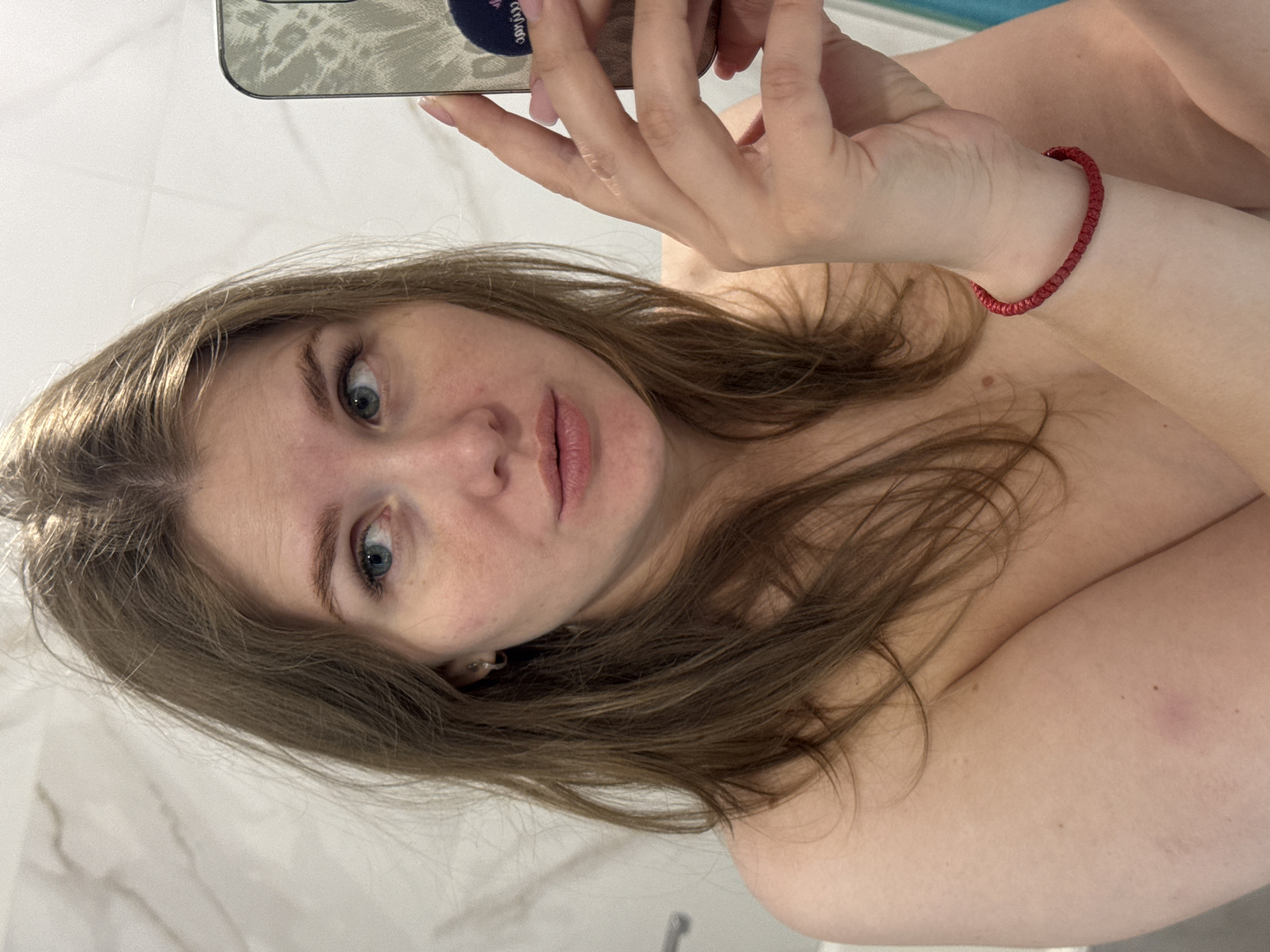 A woman is taking a selfie indoors. She has light brown hair, blue eyes, and fair skin. She is holding a smartphone with a patterned case in her left hand. A red beaded bracelet is on her left wrist. The background appears to be a bathroom with a white marble-like wall. The location is unknown, and there is no information to confirm it is Ферма, Russia.