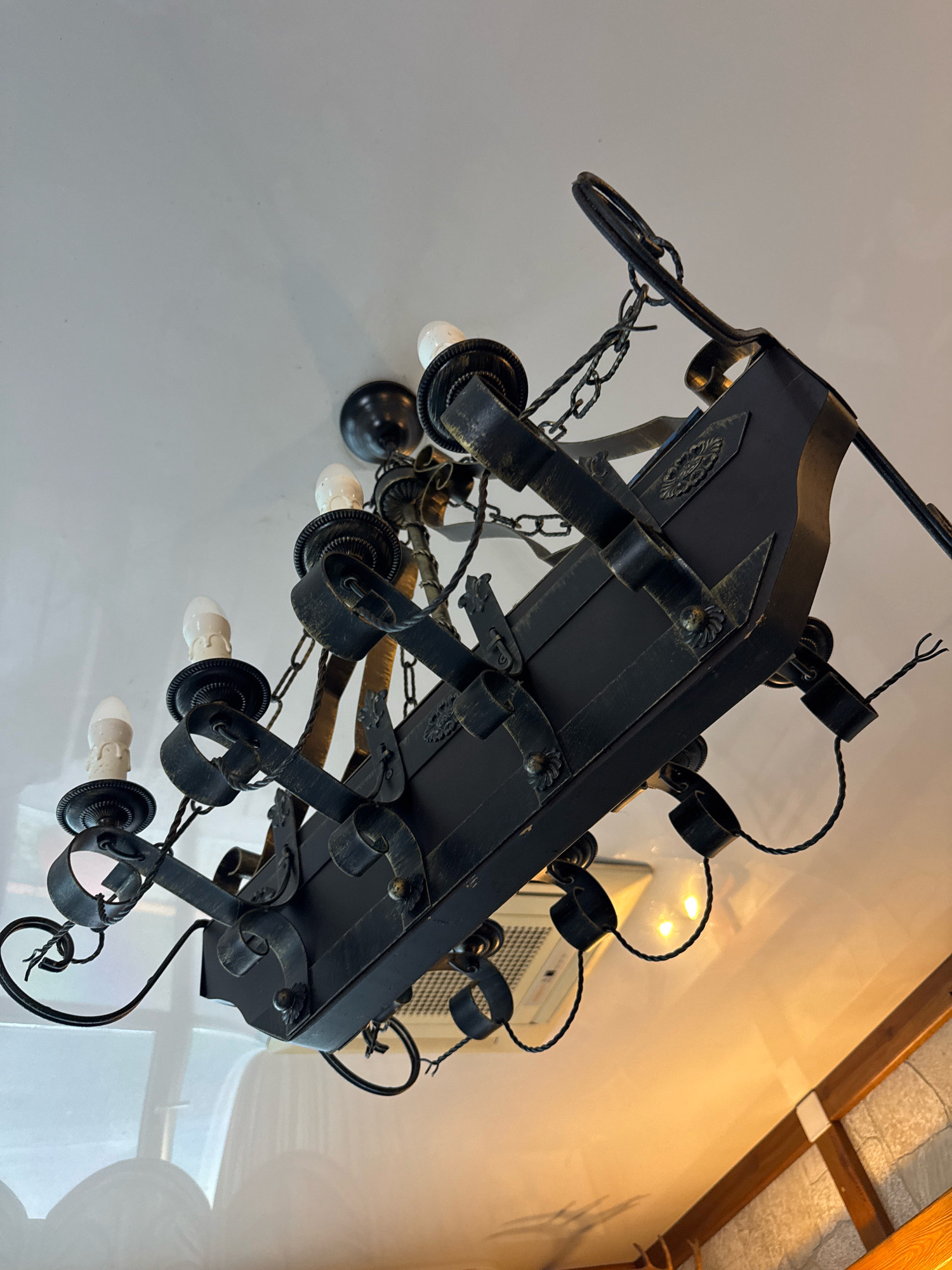 The image shows a close-up, angled view of a dark, wrought-iron chandelier suspended from a white ceiling. The chandelier has multiple candle-style light bulbs, some of which are illuminated with a warm, yellow glow. The design of the chandelier appears rustic or antique, with decorative chains, straps, and scrollwork. In the background, parts of a white ceiling and a section of wooden trim are visible. The lighting suggests an indoor setting, possibly a home or restaurant. The image does not provide any information about the city of Успенское, Russia, or any specific location within it.