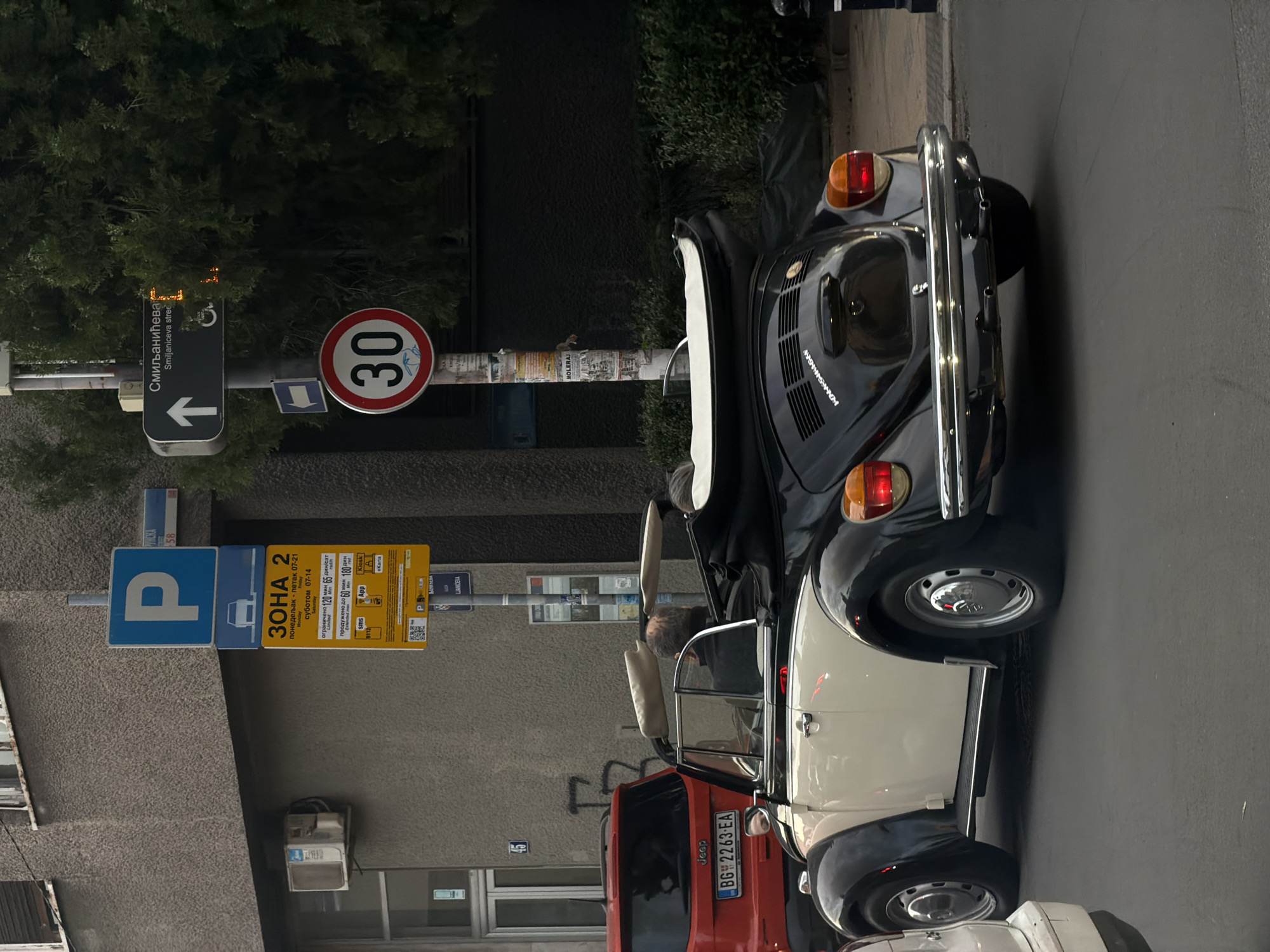 The scene is a street in Vracar Urban Municipality, Serbia, as suggested by the parking zone sign "ЗОНА 2". A vintage Volkswagen Beetle convertible is parked on the right side of the street, its top down, revealing the interior. Behind it is a red Jeep with the license plate "BG 2263-EA". Street signs on the left indicate "P" for parking, a "30" speed limit sign, and the street name "Смиљанићева (Smiljaniceva Street)" with an arrow pointing left. There's a person visible inside the Beetle. The surrounding environment includes buildings and greenery, suggesting an urban setting.
