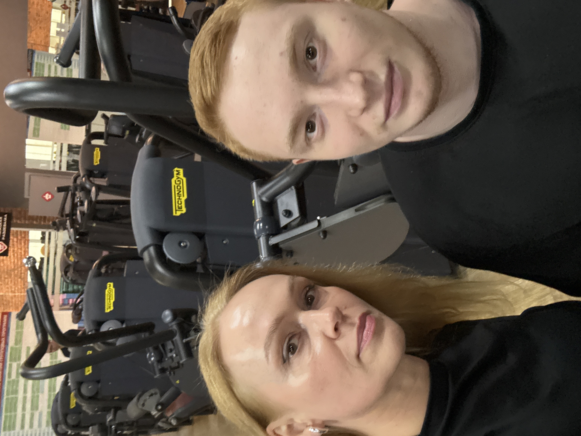 A selfie photograph shows two people, a fair-skinned male with reddish-blonde hair and a fair-skinned blonde female, likely taken indoors in a gym. Both individuals are wearing dark-colored shirts. Behind them are several stationary bikes. The brand "Technogym" is visible on the bikes. The bikes are dark-colored. Further in the background, posters are visible. The location is likely a gym in the city of Chaikovsky, Russia.