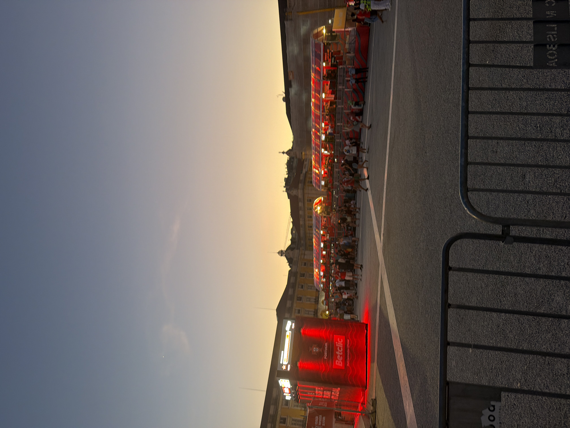The image depicts an outdoor public square in Lisbon, Portugal, at dusk. A large building with yellow facades and arched windows forms the backdrop. In the foreground, a red, illuminated kiosk with the "Betclic" logo and "PORTUGAL" branding stands prominently. To the right, a long, multi-tiered red structure, possibly a stage or promotional display, is visible with people gathered around and on it. The ground is paved with asphalt, and metal barriers line the lower edge of the frame. The sky transitions from a pale blue at the top to a warm orange hue near the horizon, indicating the setting sun. Several individuals are present in the scene, standing, walking, and observing the attractions. The overall atmosphere suggests a public event or celebration.