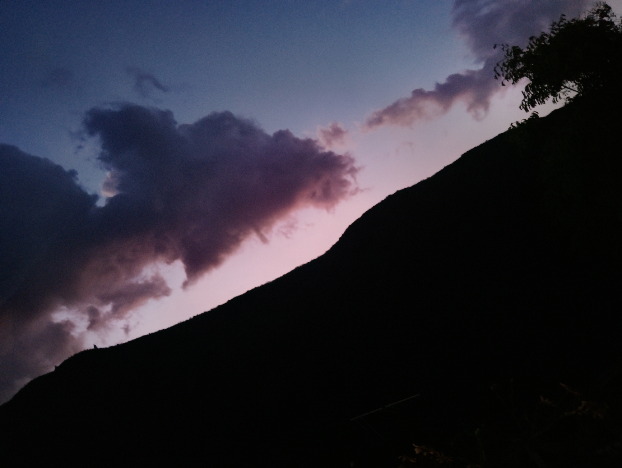 The image shows a dark silhouette of a hill against a twilight sky. The sky is transitioning from blue to pink, with patches of purple-tinged clouds scattered across it. A tree is silhouetted on the upper right side of the hill. The lower portion of the image is dark, obscuring any details on the ground. The location is Caracas, Venezuela.