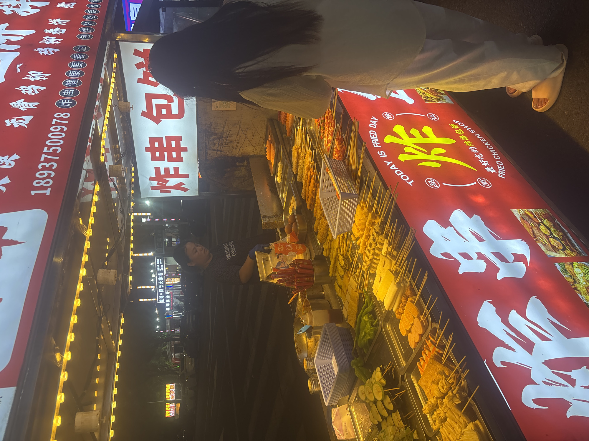 The image depicts a bustling night market scene in Suzhou Industrial Park, China, focusing on a food stall specializing in fried skewers. A woman, likely the vendor, stands behind a counter laden with various ingredients arranged in metal trays. These include an assortment of skewered items such as vegetables like green peppers and what appear to be fried zucchini slices, as well as processed foods like fish cakes, sausages, and possibly tofu products. Red chopsticks in a holder and various condiment bottles are also visible on the counter. A customer, a young woman with long dark hair, stands in front of the stall, looking at the food. Above the food display, banners and signage are prominent. One banner features Chinese characters and a phone number, while another, larger banner displays "炸串 包饼" (fried skewers, wrapped pancakes) in large red characters, and the English phrase "TODAY IS FRIED DAY" and "FRIED CHICKEN SHOP" below. The stall is illuminated by bright yellow string lights, creating a vibrant atmosphere. In the background, the night scene of the industrial park is partially visible, with other stalls and the silhouettes of cars and buildings suggesting an active commercial area. The overall impression is one of a lively, urban street food environment.
