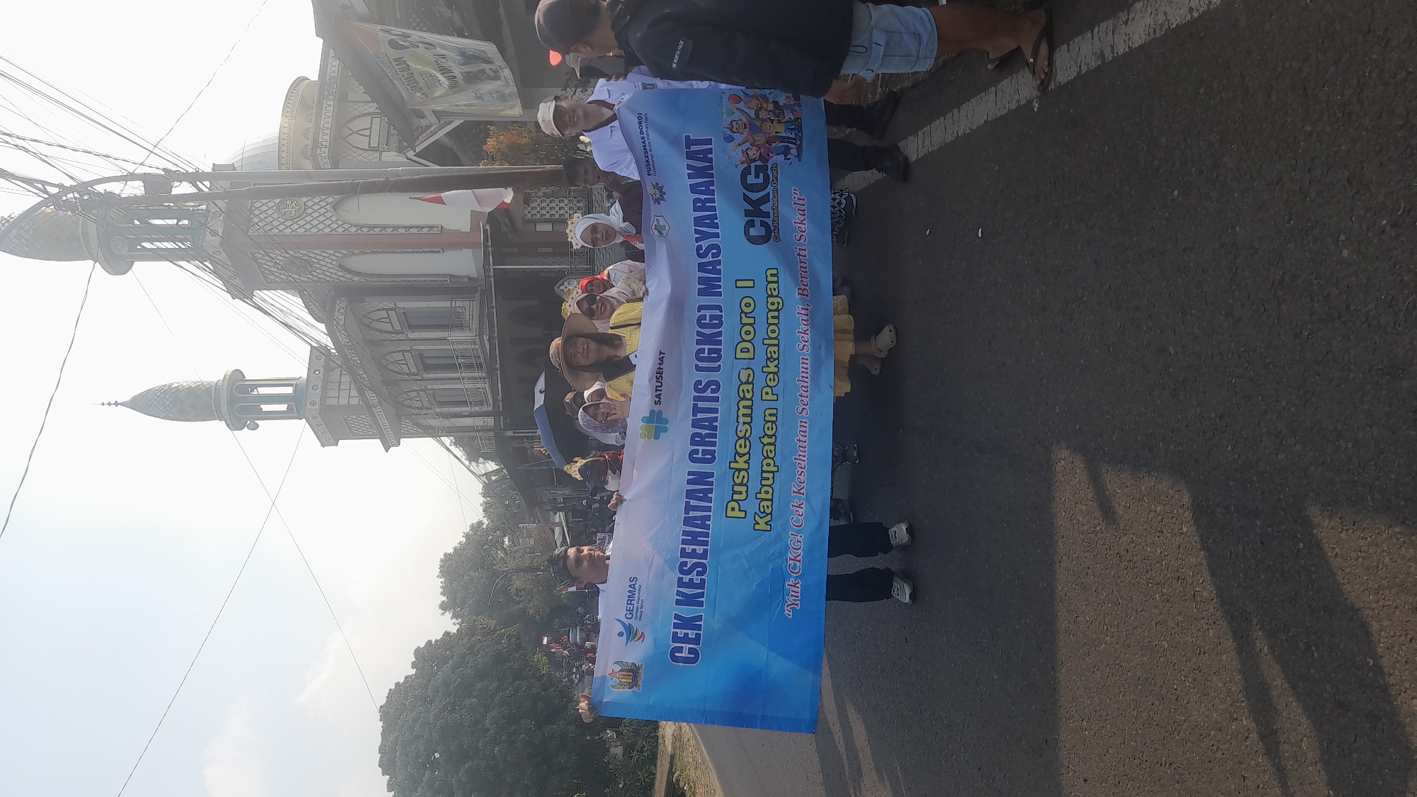 In Lemahabang, Indonesia, a group of people are participating in a parade. In the foreground, individuals hold a large blue banner that reads "CEK KESEHATAN GRATIS (GKG) MASYARAKAT PUSKESMAS DORO I KABUPATEN PEKALONGAN." The banner also features the text "Yuk CKG! Cek Kesehatan Setahun Sekali, Berarti Sekali." On the left side of the banner, there are logos for GERMAS and SATUSEHAT. Several participants are smiling and looking at the camera, including a woman in a yellow dress and a hat, and a man in a white lab coat. In the background, a mosque with a dome and minaret is visible, along with trees and buildings. The parade appears to be taking place on a paved road.
