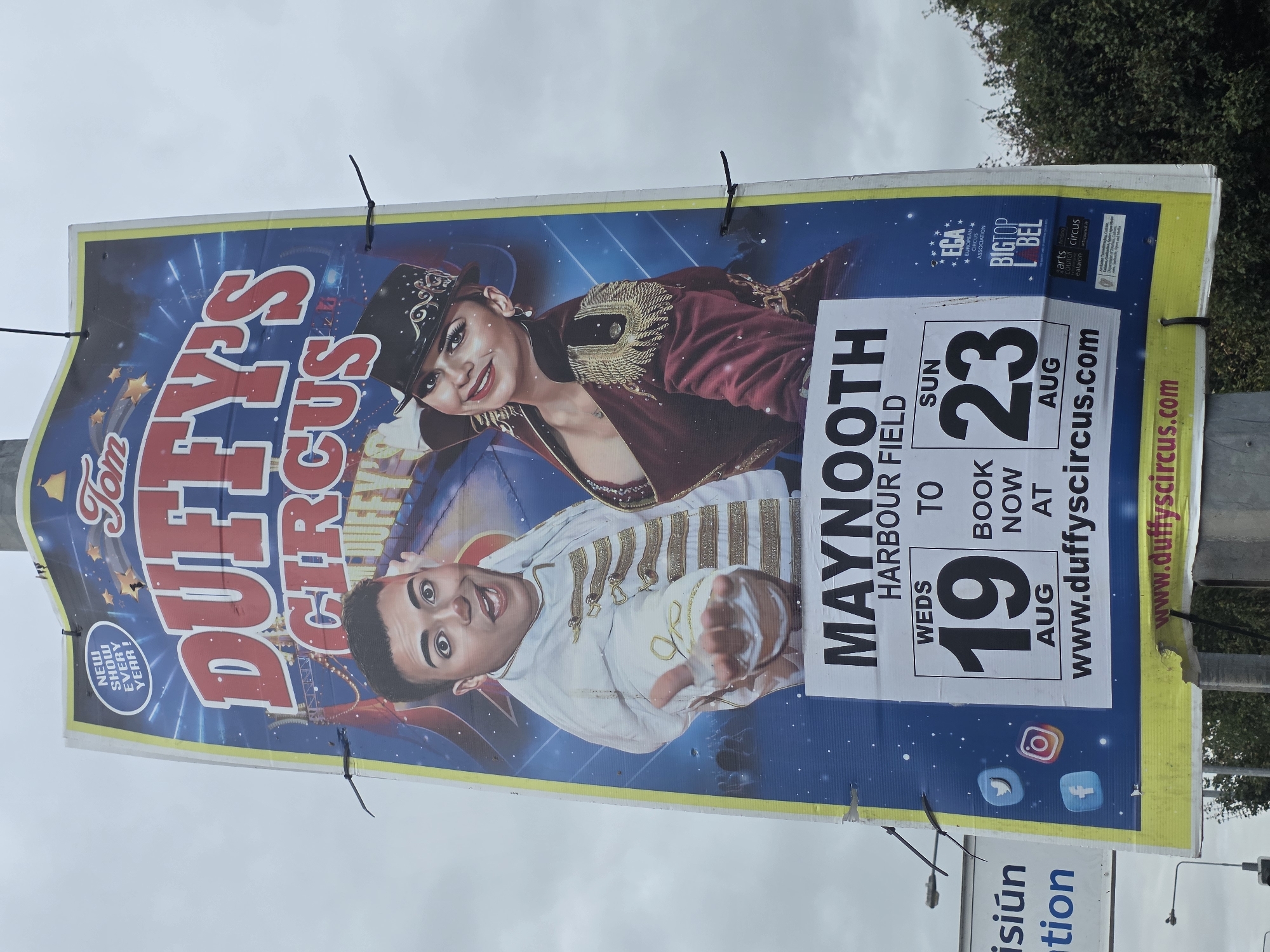 This is a photograph of a large billboard advertisement for Tom Duffy's Circus, located in Maynooth, Ireland. The billboard is predominantly blue with a starfield background and features two performers in circus attire. A male performer in a white and gold jacket is on the left, and a female performer in a burgundy and gold outfit with a hat is on the right.
Below the performers, the text "MAYNOOTH HARBOUR FIELD" is prominently displayed. Further down, dates are advertised: "WEDS 19 AUG" and "SUN 23 AUG", with "BOOK NOW AT" between them. A website address, "www.duffyscircus.com", is visible at the bottom.
Social media icons for Facebook, Instagram, and Twitter are present on the left side of the billboard. On the right, there are logos for "ECA EUROPEAN CIRCUS ASSOCIATION", "BIG TOP LABEL", and various arts council logos.
The billboard is affixed to a pole and appears to be outdoors, with a cloudy sky overhead. The surrounding environment shows glimpses of trees and other signs, including one that reads "ision" and "tion" in blue text, suggesting this is a public area with other advertisements or information. The billboard itself shows some signs of wear and tear, with small holes and some peeling.