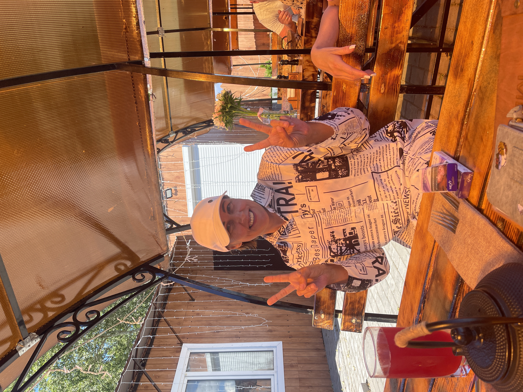 A woman wearing a white baseball cap and a shirt with a newspaper print sits at a wooden table outdoors under a translucent amber canopy. She is smiling and holding up two fingers in a peace sign with each hand. In front of her is a glass of red liquid, a black teapot, a folded napkin with a fork, and a pack of cigarettes. The background shows a wooden building and trees. The scene is likely a cafe or restaurant in Belozersk, Russia.