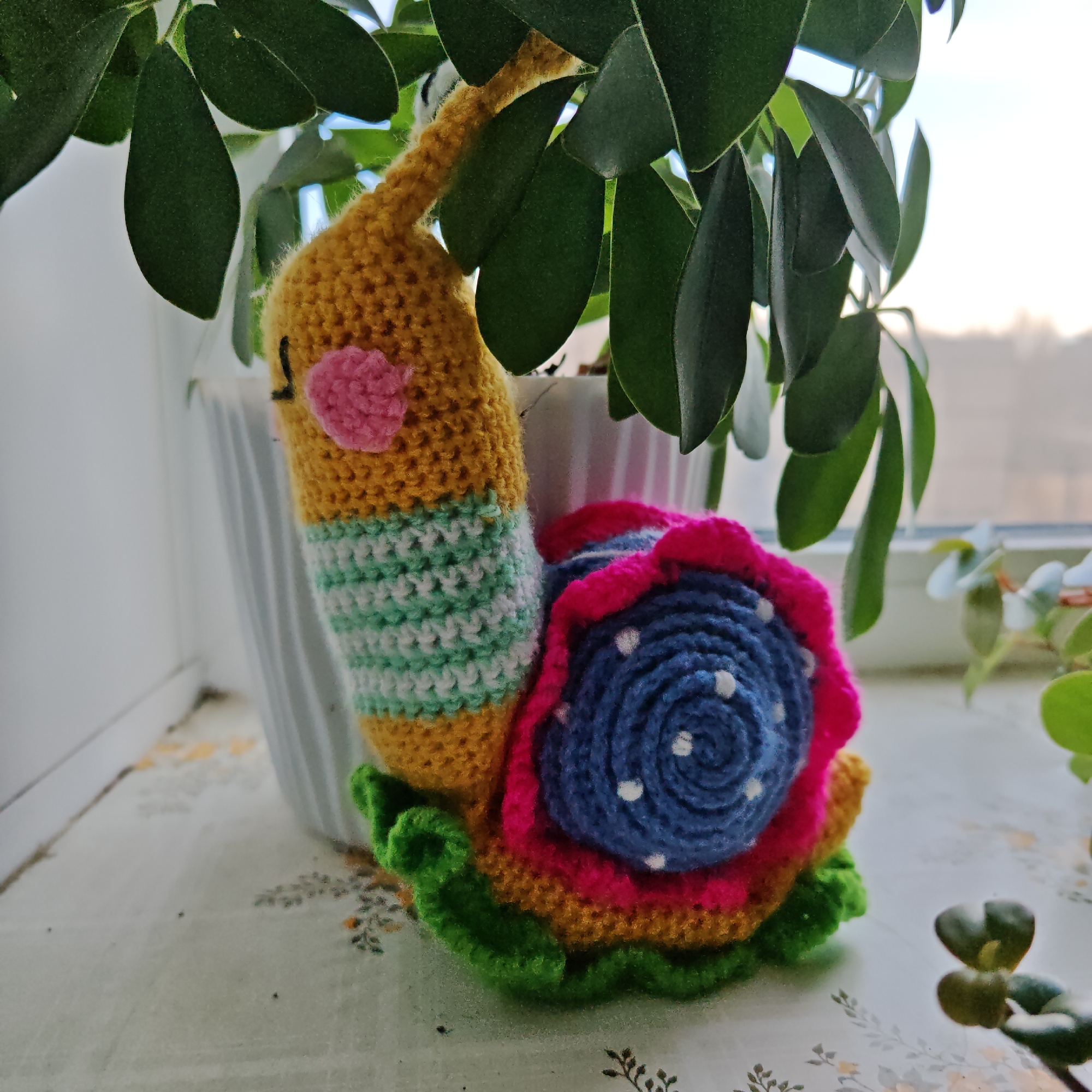 A crocheted snail toy is positioned on a surface next to a houseplant in a white pot. The snail is multicolored, featuring a yellow body with green and white stripes, a pink cheek, and a shell composed of green, yellow, pink, and blue layers. The blue layer of the shell has white dots. The plant has small, round, green leaves. The setting appears to be indoors near a window. It is impossible to pinpoint the precise location as Rostov, Russia based on the image.