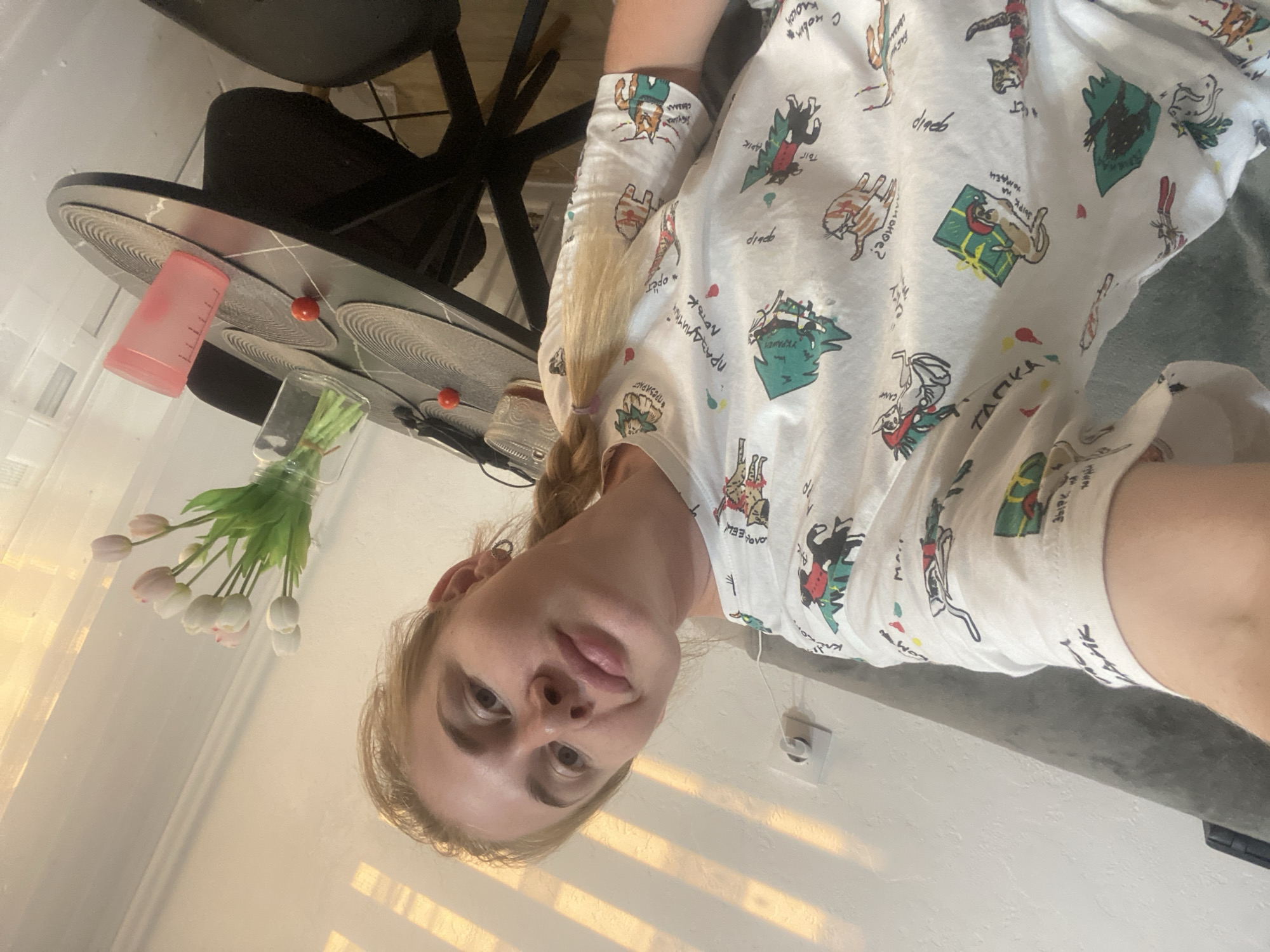 A blonde woman with her hair in a braid is taking a selfie in a brightly lit room. She is wearing a white t-shirt with a cartoon print of cats and Christmas trees. The room appears to be a living room or dining area, with a dark gray sofa visible in the background. A black round table is in the foreground, set with placemats, a glass vase with tulips, and two cherry tomatoes. A pink cup and a glass with a dark liquid are also on the table. The lighting suggests it is daytime, with sunlight filtering through blinds onto the wall behind the woman. The location of Novaya Usman', Russia is not evident from the image.