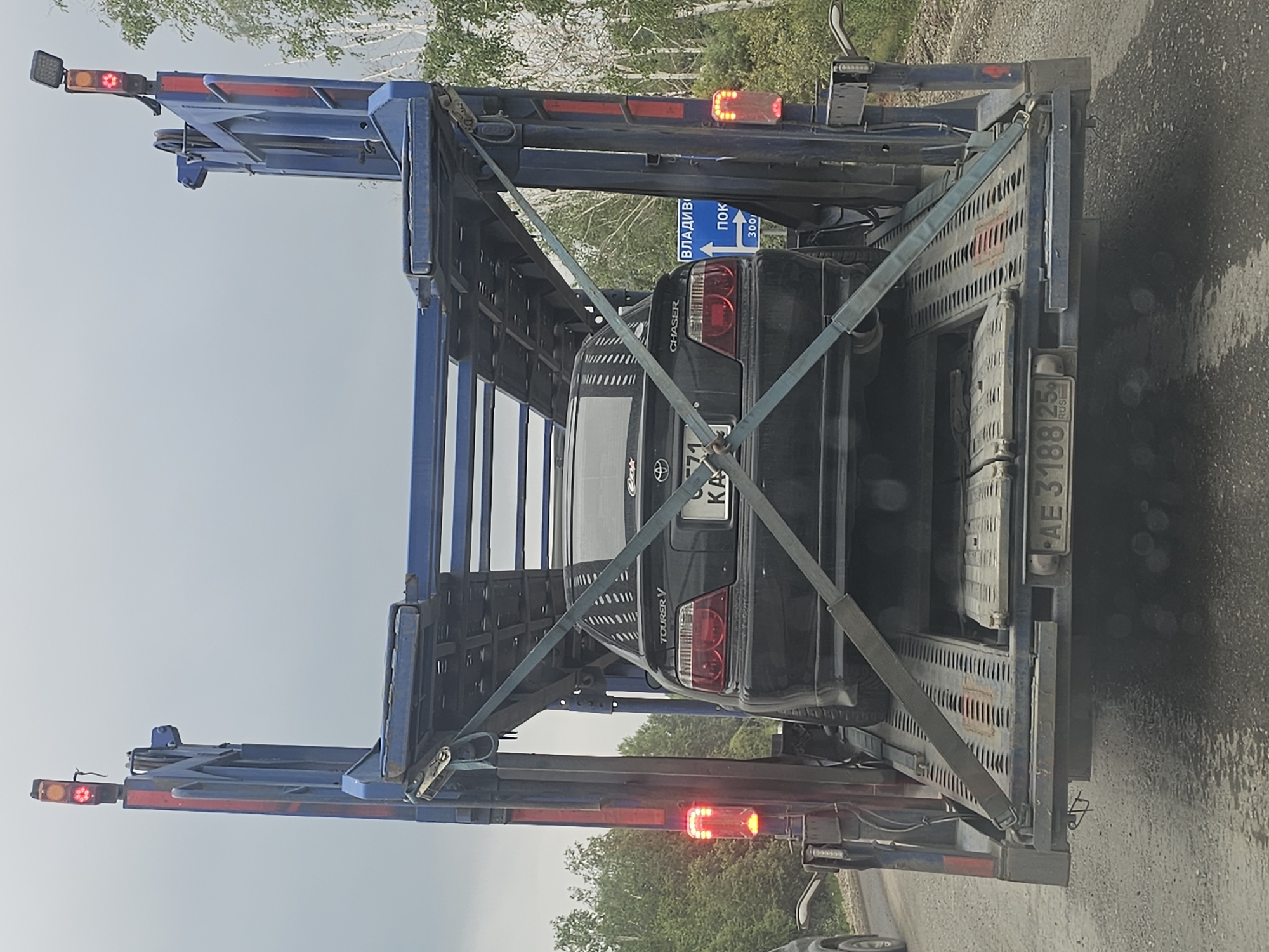 An overhead view of a black Toyota Chaser Tourer V being transported on a car carrier trailer is captured on a cloudy day. The vehicle, with license plate "K 71 KA," is secured to the trailer with blue straps. The trailer itself is blue and has red brake lights visible on its rear. A blue road sign with white text and arrows is attached to the right side of the trailer, indicating directions to "ВЛАДИВ" and "ПОК," with a distance of "300." The trailer's license plate reads "AE 3188 25 RUS." The environment appears to be a rural roadside with trees and a gray, wet road surface, suggesting it might be located near Lesopil'noe, Russia.