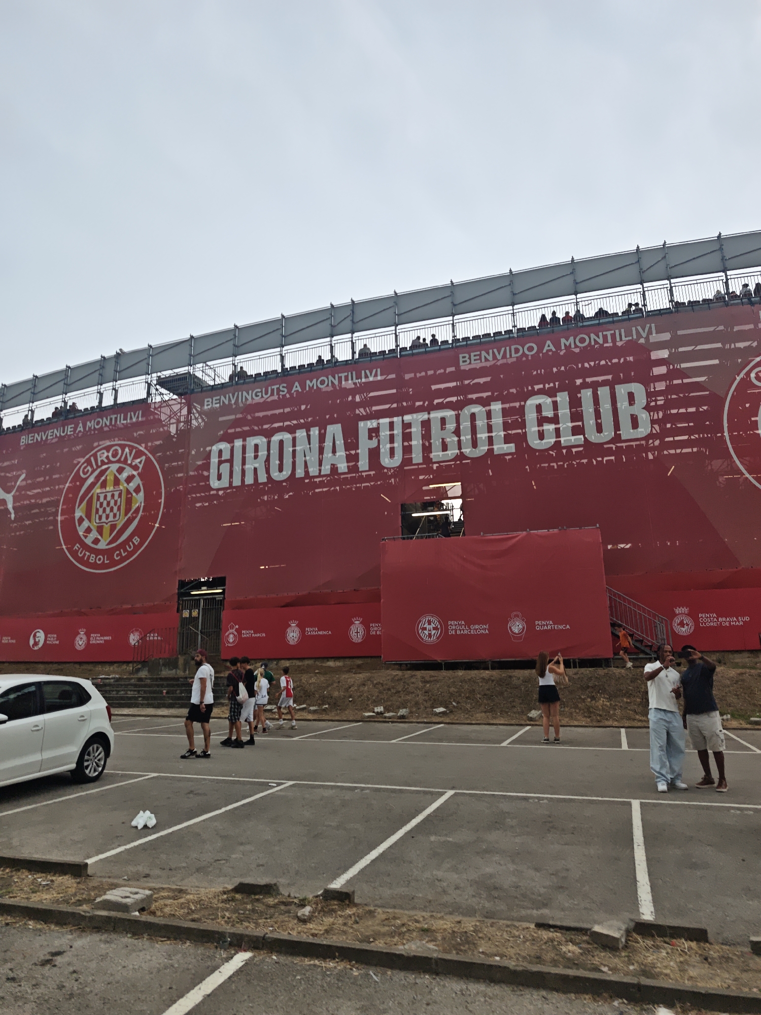A wide shot captures a large red banner draped across the exterior of a stadium. The banner displays the text "GIRONA FUTBOL CLUB" in large white letters, along with the Girona Fútbol Club crest, and the slogans "BIENVINGUTS A MONTILIVI" and "BENVIDO A MONTILIVI". Several people are visible in the foreground, standing in a parking lot. To the left, a white car is parked. A few individuals are walking or standing in the parking lot, with some appearing to be taking pictures. The overall scene suggests an event or gathering is taking place at the stadium.