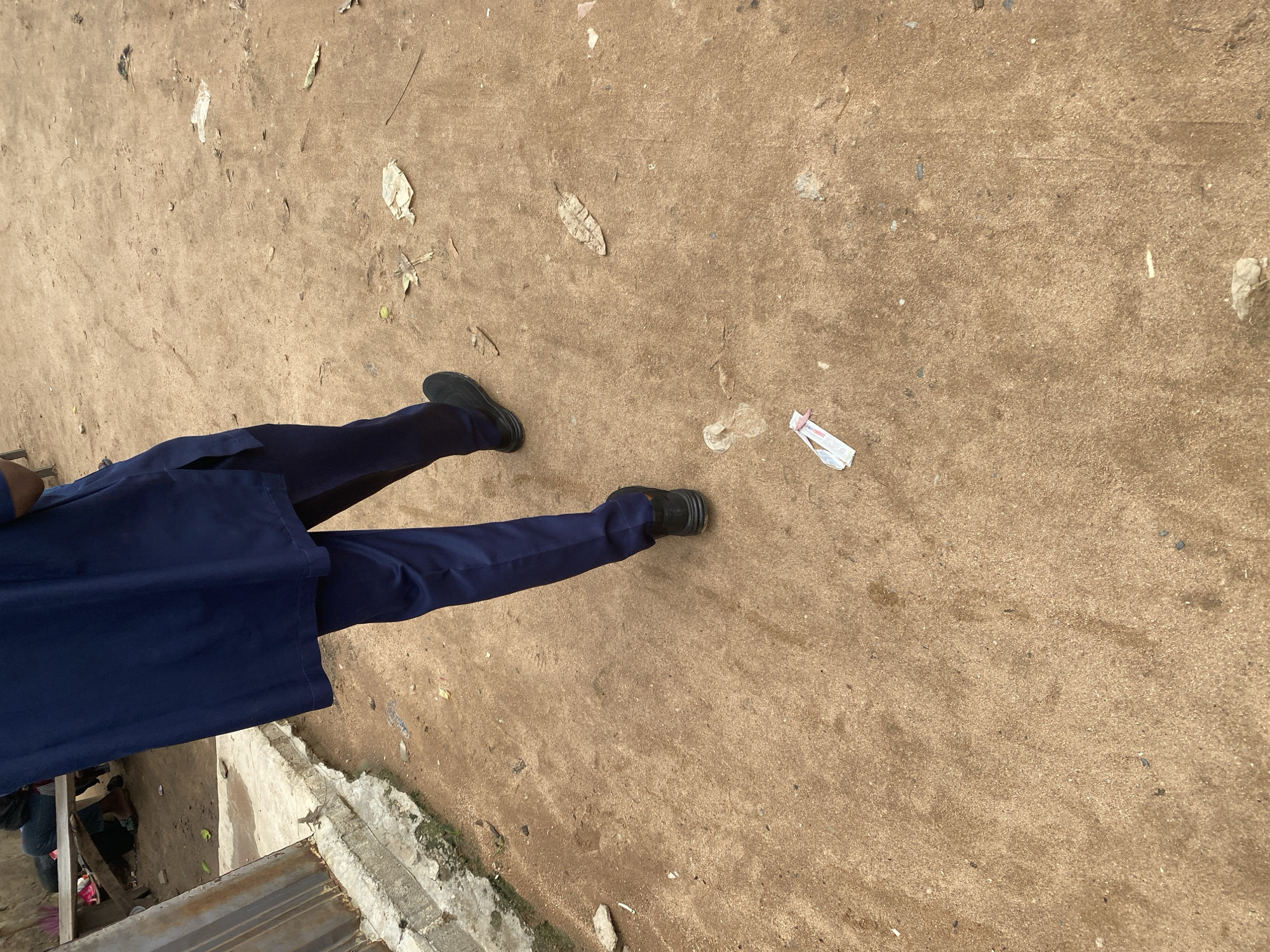The image shows the lower half of a person standing on a dirt ground. The person is wearing a navy blue uniform, which includes a jacket and trousers. They are also wearing black shoes. The ground is a mix of sand and dirt, with some dry leaves and small pieces of litter scattered across it. In the upper right portion of the image, there is a crumpled piece of white paper or plastic with pink ties. The scene appears to be outdoors, possibly in a rural or undeveloped area. The lighting suggests it is daytime. There is no visible text in the image. The location is Jalingo, Nigeria.