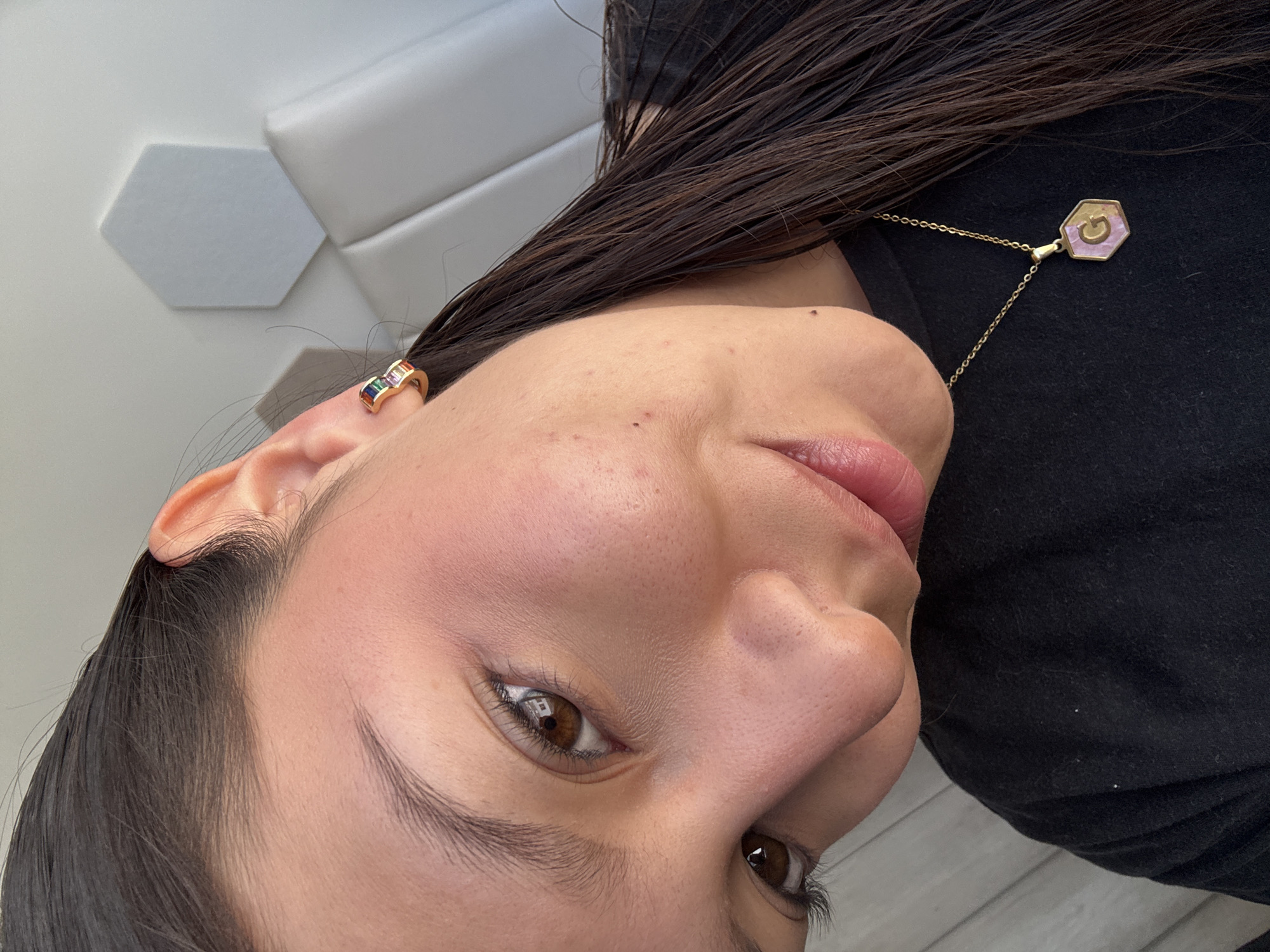 A medium-close-up, angled view focuses on a woman wearing jewelry. She has dark brown hair and brown eyes. She wears a small gold hoop earring with colored stones in her left ear. A gold necklace with a hexagonal pendant is visible near her neck. She is wearing a black top. A white, geometric wall panel is in the blurred background. The location is not identifiable.