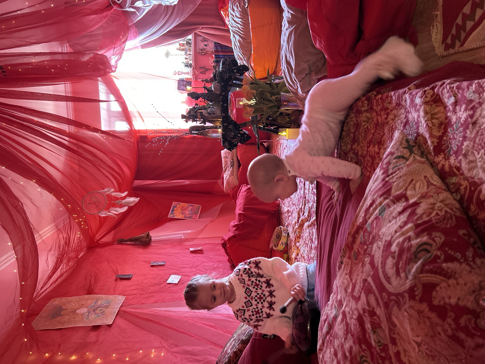 Two young children are in an indoor space decorated with red drapery and string lights. The younger child, wearing white pajamas, is crawling or climbing onto a red and brown patterned bed. The older child, wearing a white sweater with a red and black pattern, stands near the bed holding a small drum and stick. Various objects, including a dream catcher, tarot cards, plants, candles, and decorative items, are arranged within the space. The location is in a room decorated with red drapery and string lights. The specific city of Balashikha in Russia is not evident.