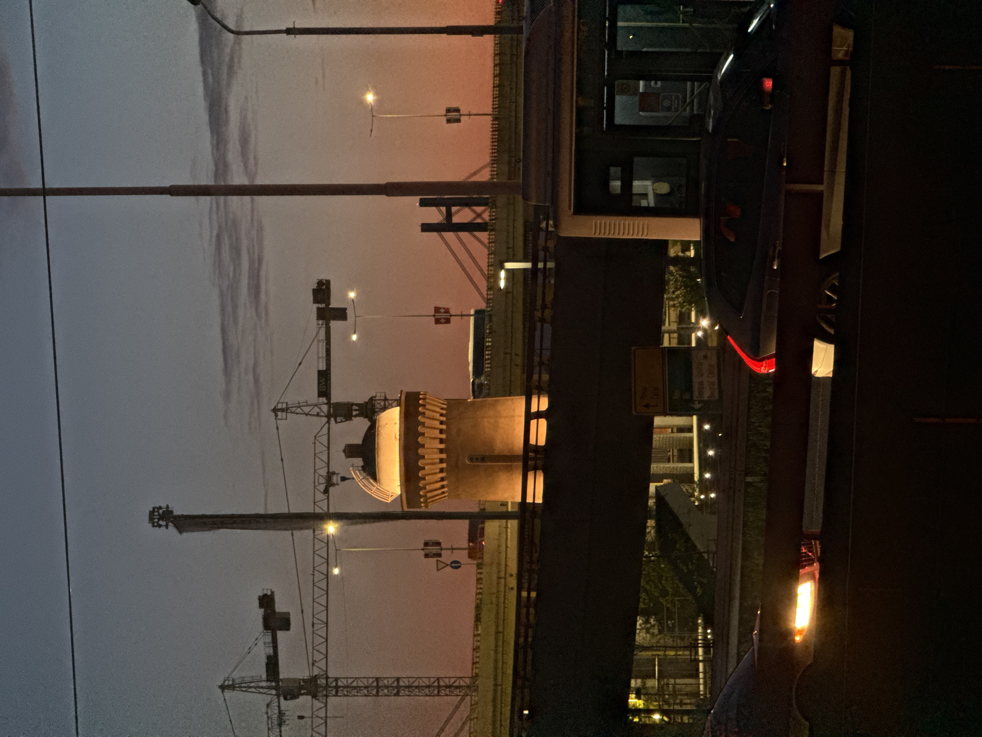 An urban landscape at either dawn or dusk is depicted, characterized by a dramatic sky transitioning from a dark grey-blue at the top to a warm orange-pink closer to the horizon. In the middle ground, a prominent, light-colored cylindrical tower is centrally positioned, illuminated from below by warm artificial lights that highlight its ribbed or crenellated upper section.
The scene suggests ongoing development with multiple large construction cranes, featuring distinctive lattice structures, visible across the left and central portions of the frame. Utility poles and power lines extend across the upper part of the sky. To the right, a dark, elevated structure, likely part of a bridge or overpass, casts a deep shadow. Bright red taillights and an orange turn signal from a partially visible vehicle pierce the darkness in the foreground, indicating active traffic. Streetlights illuminate various sections of the environment.
No specific human activity is discernible, but the illuminated tower, streetlights, and vehicle suggest an active urban environment. A faint "BW" logo is visible on one of the cranes. The image captures a blend of architectural elements and industrial infrastructure under the transitional light of daybreak or twilight in Belgrade, Serbia.